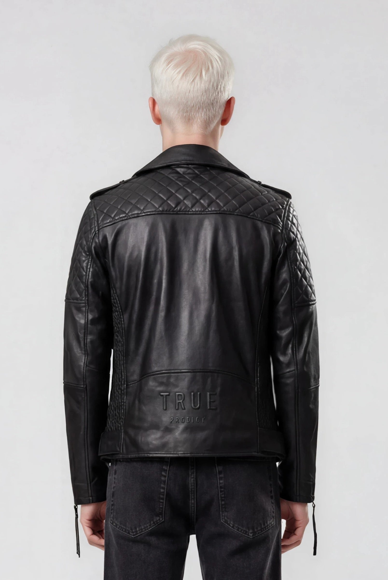 Eine Person mit kurzen, platinblonden Haaren ist von hinten zu sehen. Sie trägt die trueprodigy Biker Lederjacke Herren "Simba" mit Gürtel und schwarze Jeans und steht vor einem schlichten hellgrauen Hintergrund.