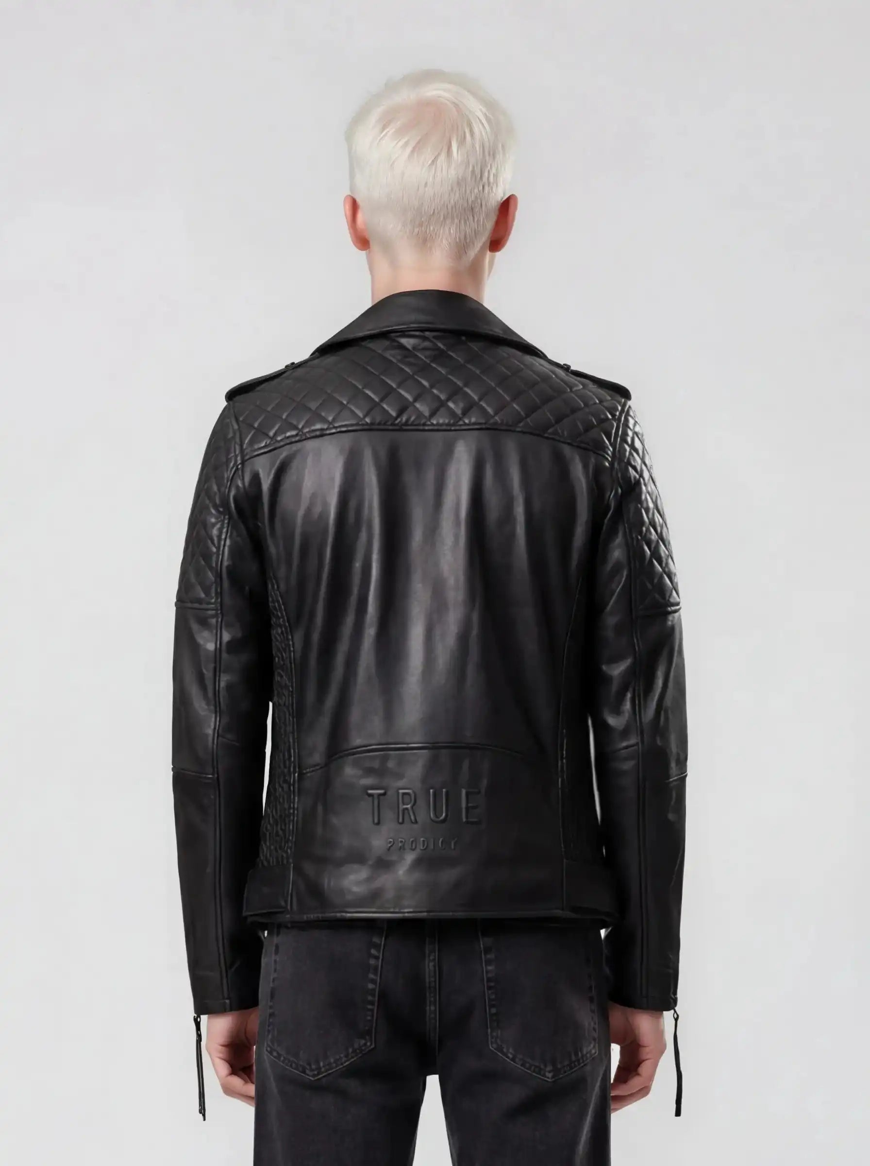 Eine Person mit kurzen, platinblonden Haaren ist von hinten zu sehen. Sie trägt die trueprodigy Biker Lederjacke Herren "Simba" mit Gürtel und schwarze Jeans und steht vor einem schlichten hellgrauen Hintergrund.