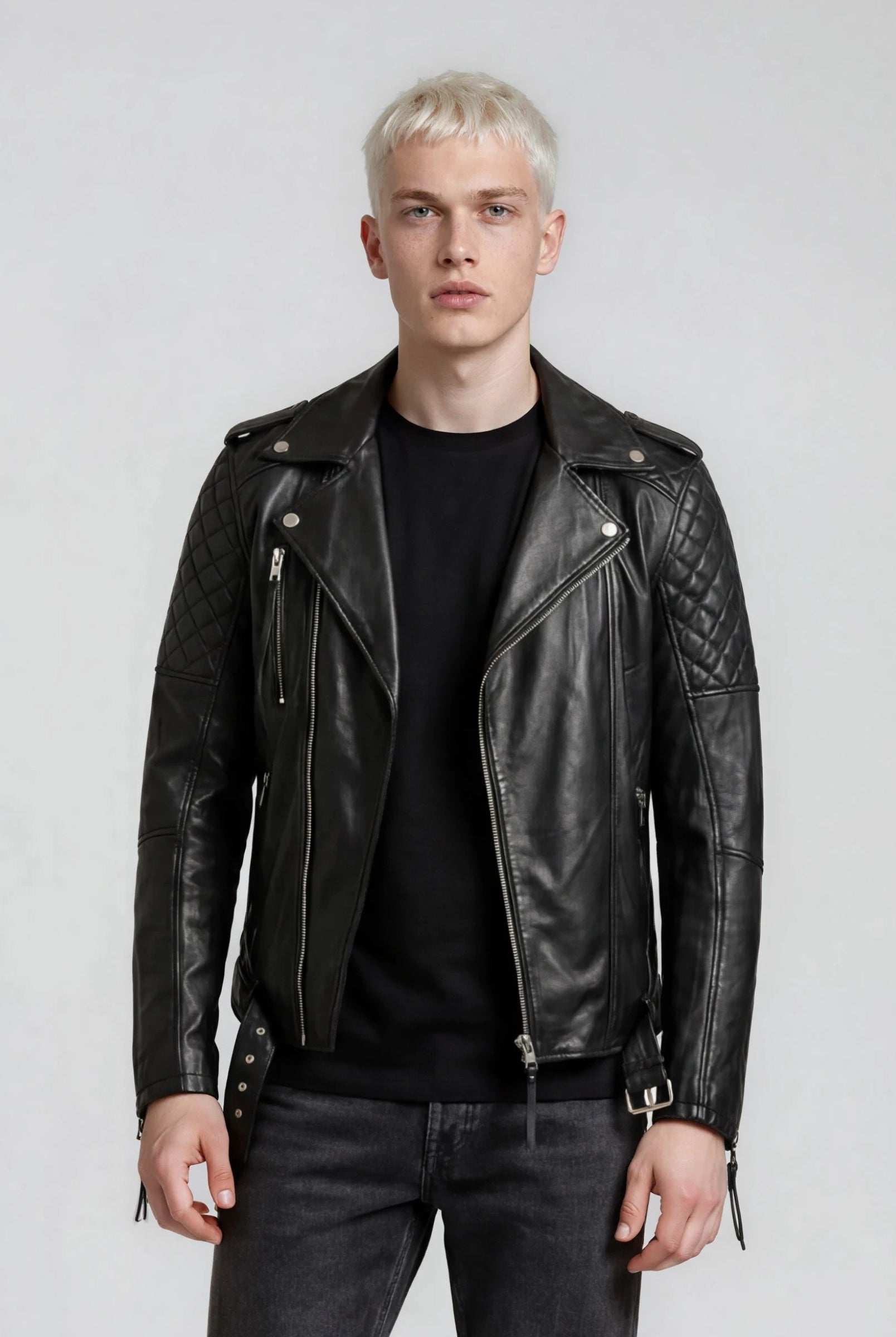 Eine Person mit kurzen blonden Haaren trägt eine trueprodigy Biker Lederjacke Herren "Simba" mit Gürtel über einem schwarzen Hemd und einer dunklen Jeans und steht vor einem schlichten hellgrauen Hintergrund.