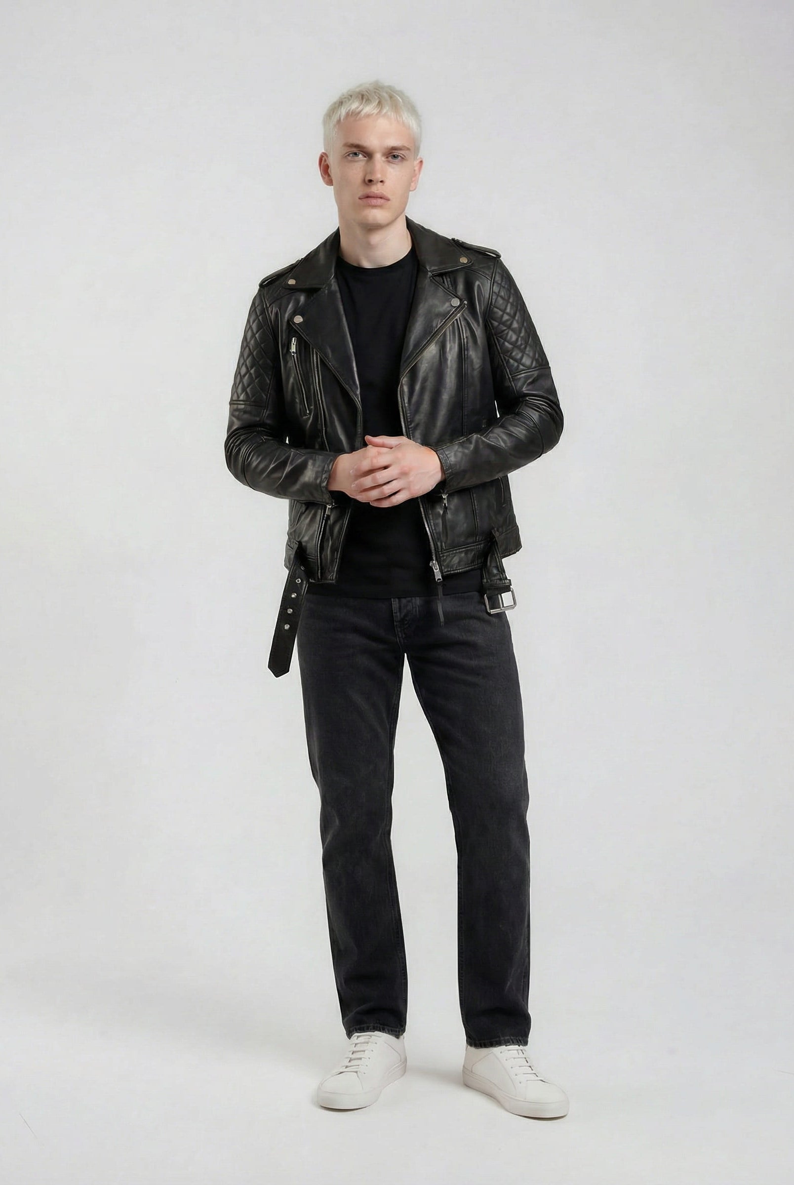 Eine Person mit kurzen, hellen Haaren steht vor einem schlichten Hintergrund und trägt eine trueprodigy Biker Lederjacke Herren "Simba" mit Gürtel, ein schwarzes Hemd, dunkle Jeans und weiße Turnschuhe. Ihre Hände sind auf Hüfthöhe ineinander verschränkt.
