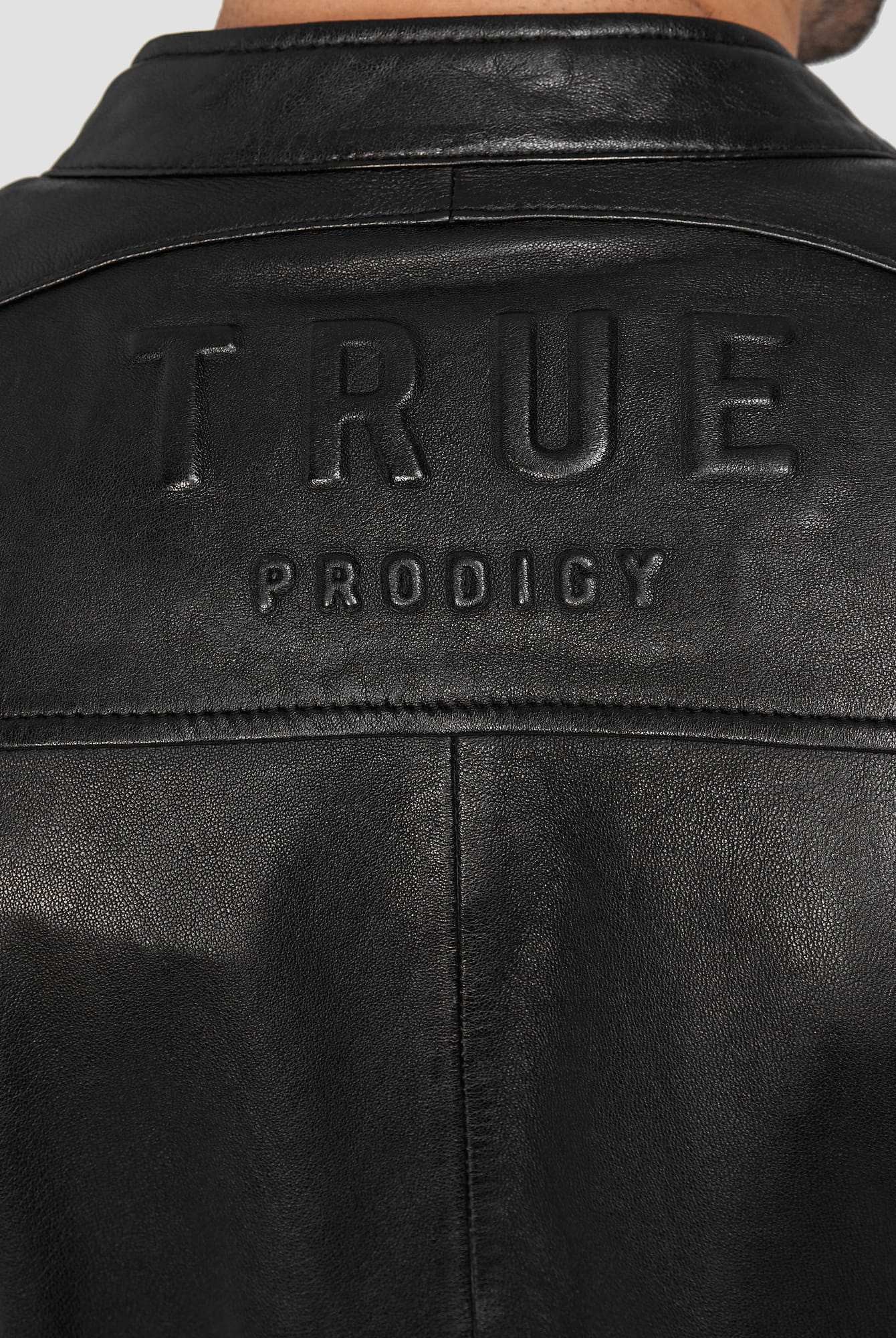 Nahaufnahme der Rückseite einer schwarzen trueprodigy Franky Lederjacke mit Stehkragen mit der Prägung TRUEPRODIGY in Großbuchstaben auf dem oberen Teil.