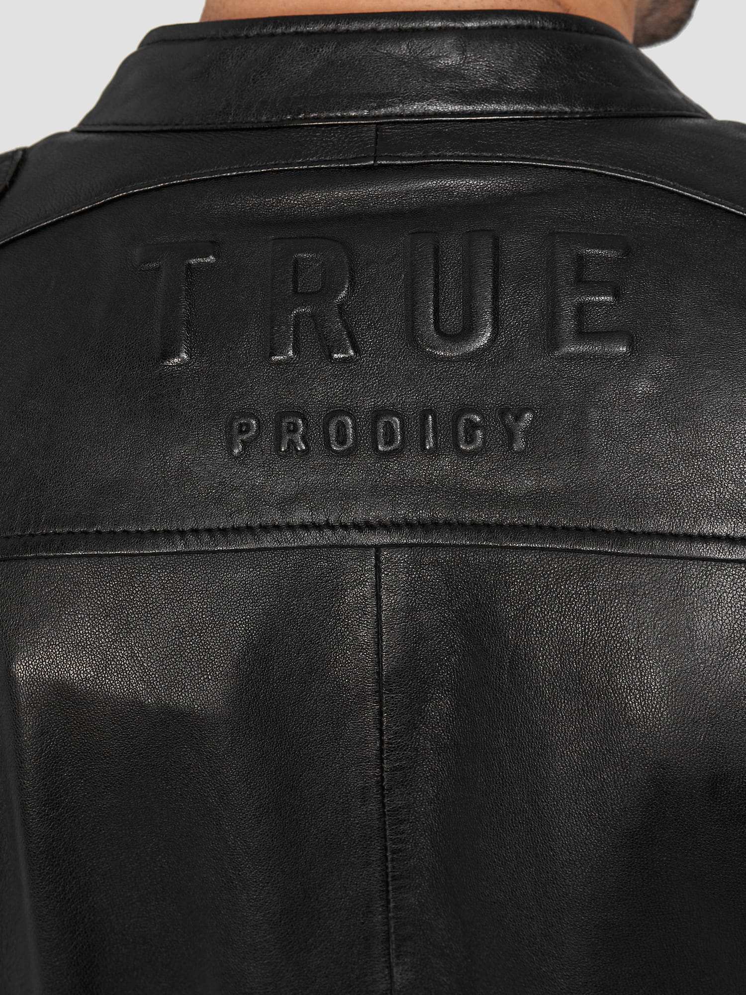 Nahaufnahme der Rückseite einer schwarzen trueprodigy Franky Lederjacke mit Stehkragen mit der Prägung TRUEPRODIGY in Großbuchstaben auf dem oberen Teil.