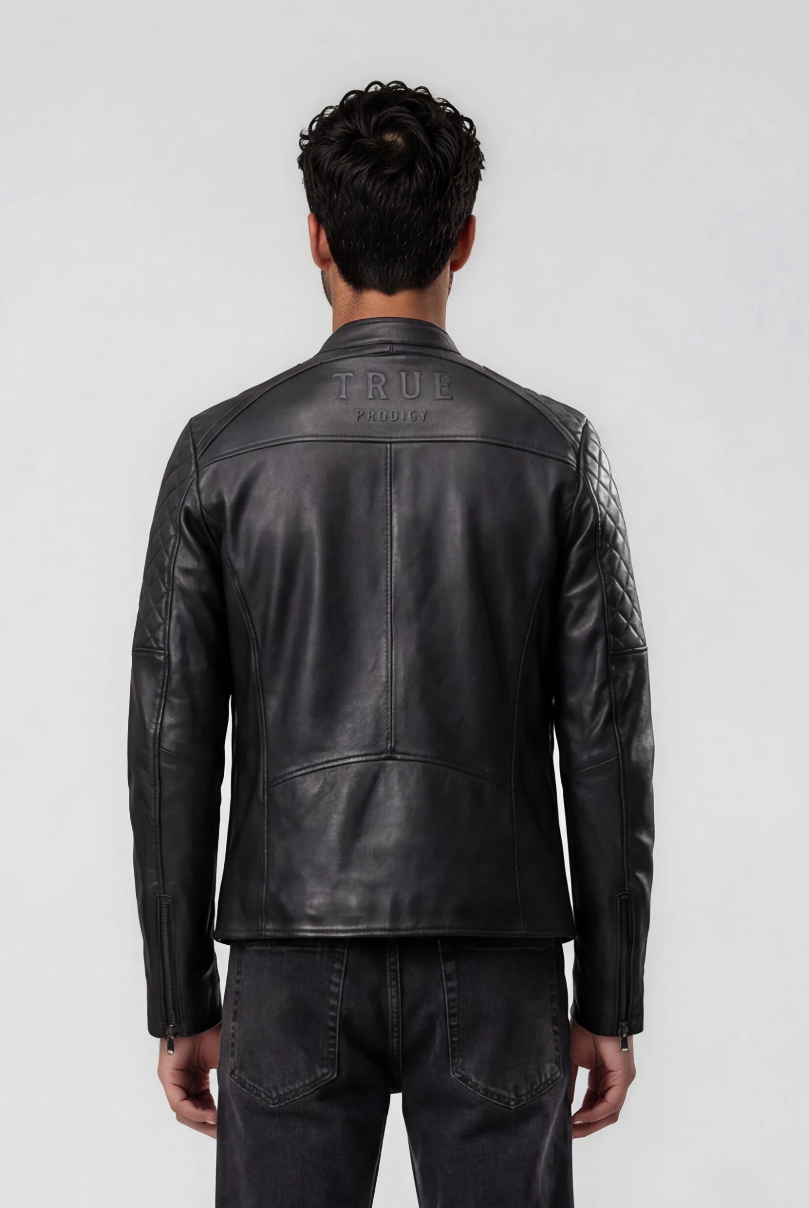Ein Mann blickt in einer schwarzen trueprodigy Racing Lederjacke Herren "Franky" mit Rautensteppung und dunkler Jeans von der Kamera weg. Auf dem oberen Rücken befindet sich ein auffälliges Branding. Der Hintergrund ist einfarbig, hellgrau.