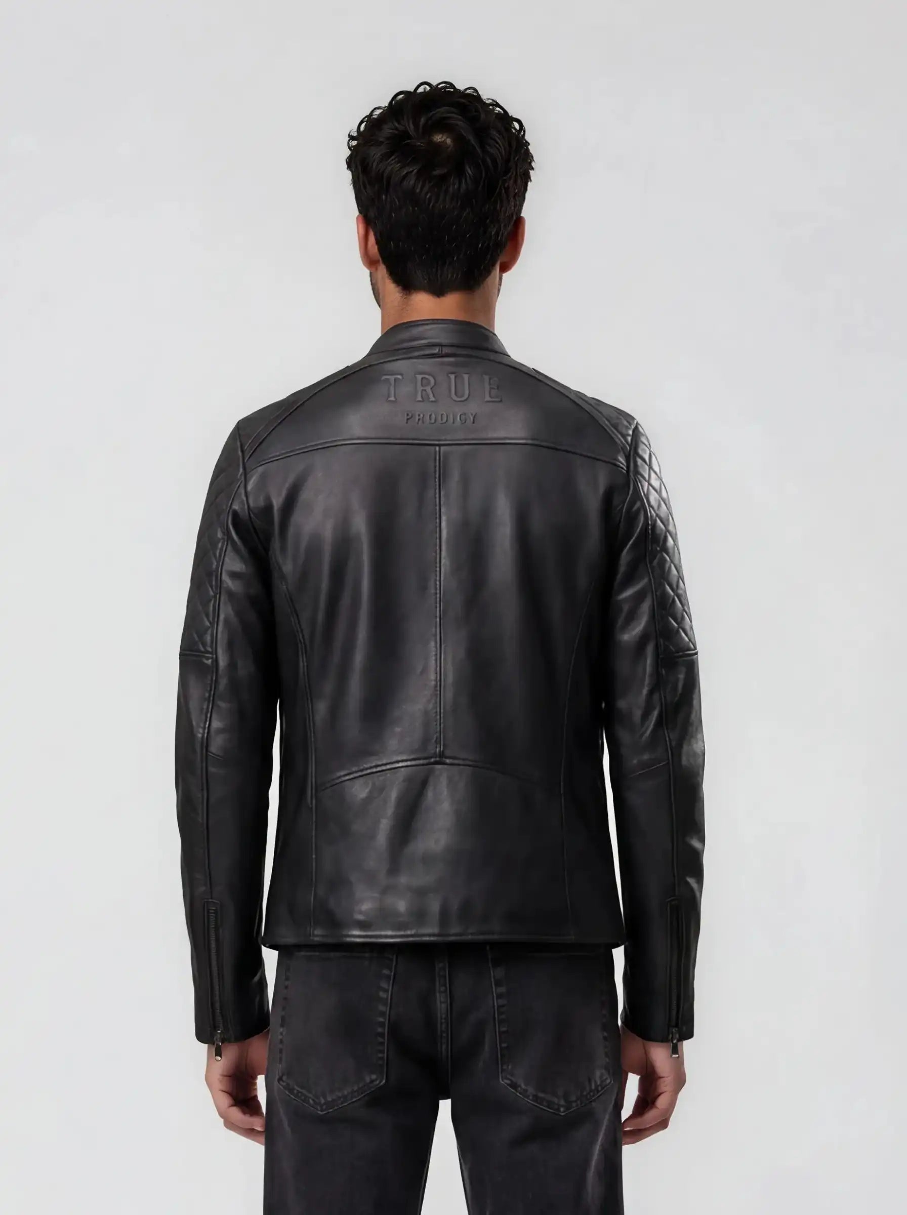 Ein Mann blickt in einer schwarzen trueprodigy Racing Lederjacke Herren "Franky" mit Rautensteppung und dunkler Jeans von der Kamera weg. Auf dem oberen Rücken befindet sich ein auffälliges Branding. Der Hintergrund ist einfarbig, hellgrau.