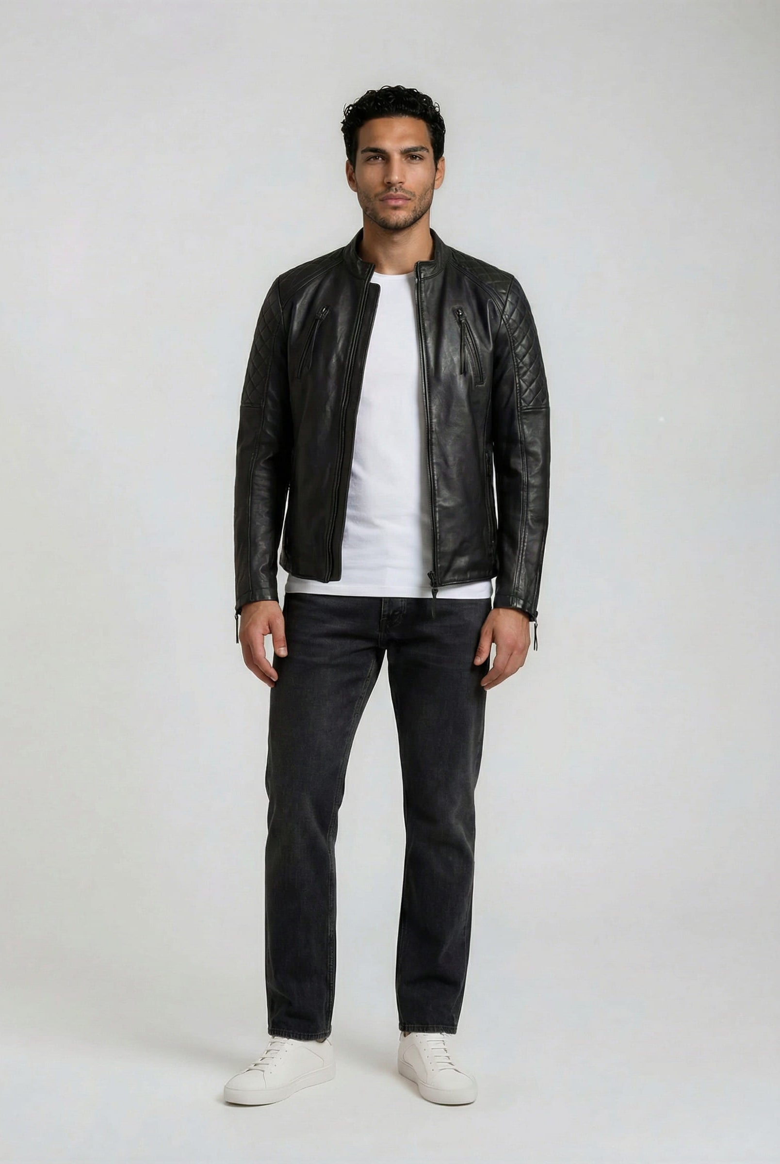 Ein Mann schaut nach vorne und trägt die trueprodigy Racing Lederjacke Herren "Franky" mit Rautensteppung, gestylt mit weißem T-Shirt, dunkler Jeans und weißen Turnschuhen vor einem schlichten hellen Hintergrund.