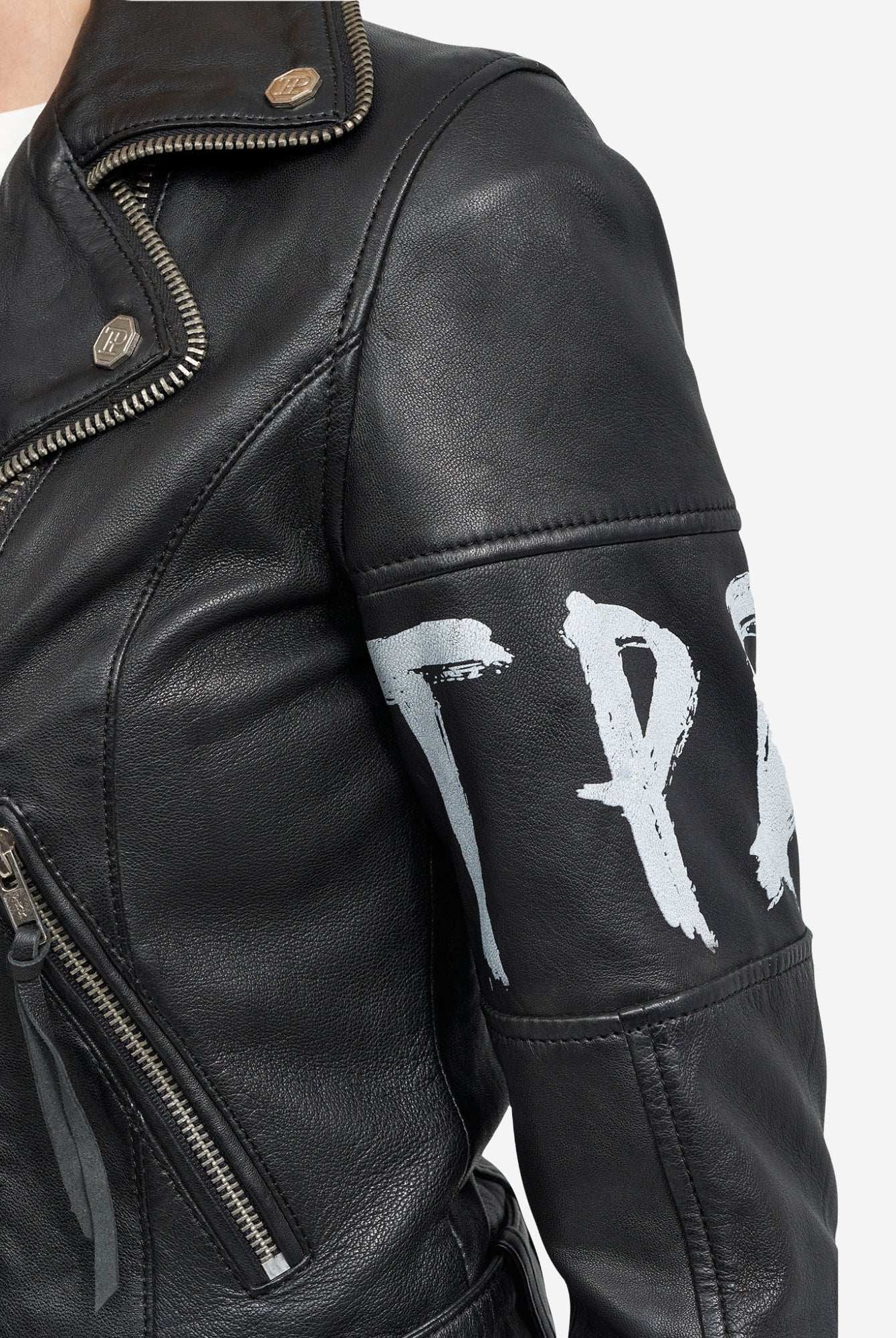Eine Nahaufnahme der trueprodigy Laila Lederjacke mit Druck zeigt schwarzes Leder mit silbernen Reißverschlüssen, Druckknöpfen und teilweise sichtbaren weiß aufgemalten Buchstaben auf dem oberen Ärmel. Der Hintergrund ist einfarbig weiß.