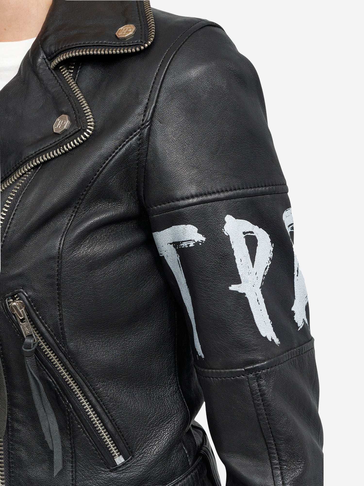 Eine Nahaufnahme der trueprodigy Laila Lederjacke mit Druck zeigt schwarzes Leder mit silbernen Reißverschlüssen, Druckknöpfen und teilweise sichtbaren weiß aufgemalten Buchstaben auf dem oberen Ärmel. Der Hintergrund ist einfarbig weiß.
