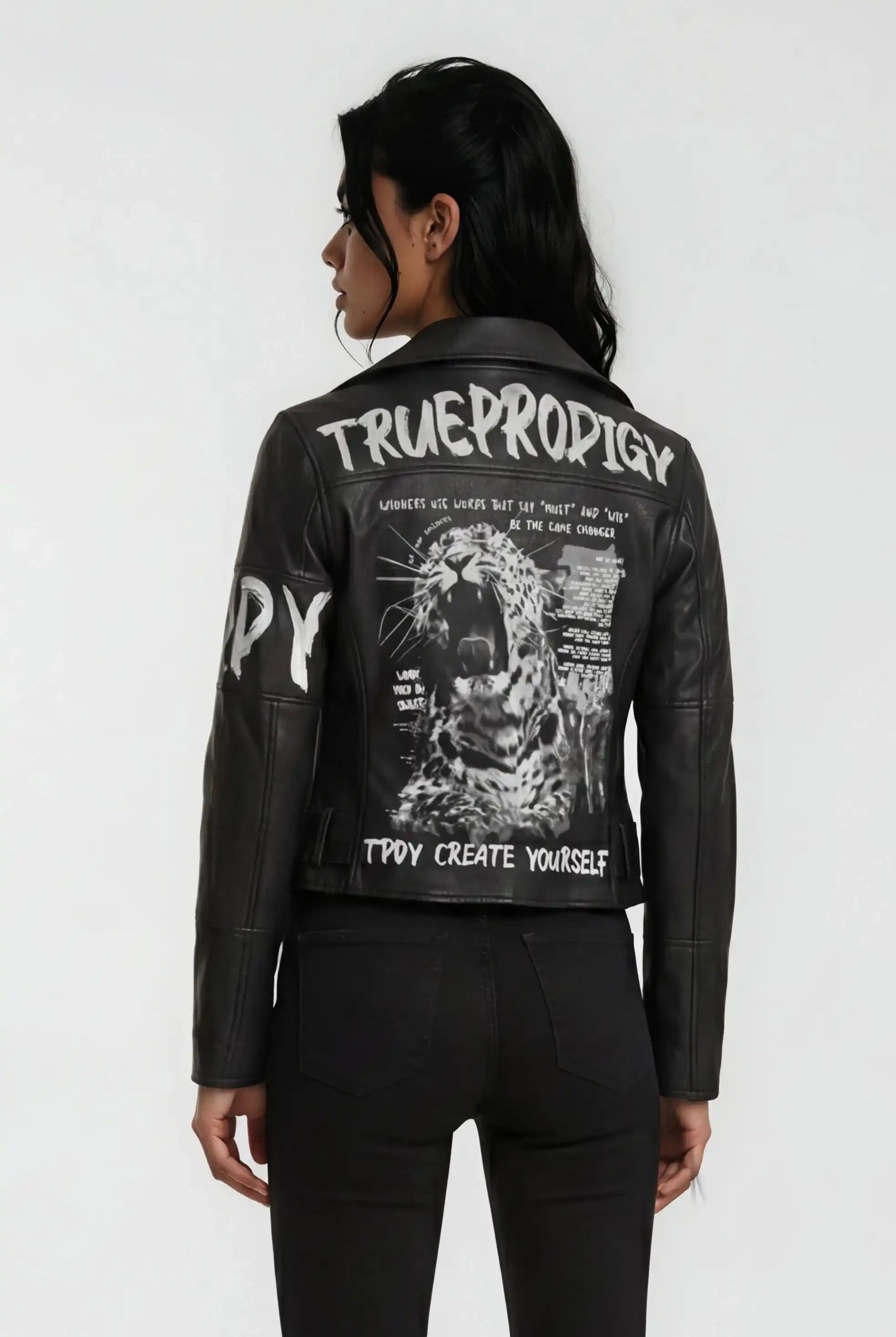 Eine Person mit langen dunklen Haaren, die die trueprodigy Lederjacke mit Druck mit weißen grafischen Motiven, einem brüllenden Leoparden und den Worten TRUEPRODIGY und CREATE YOURSELF auf der Rückseite trägt, steht mit dem Gesicht zur Wand.