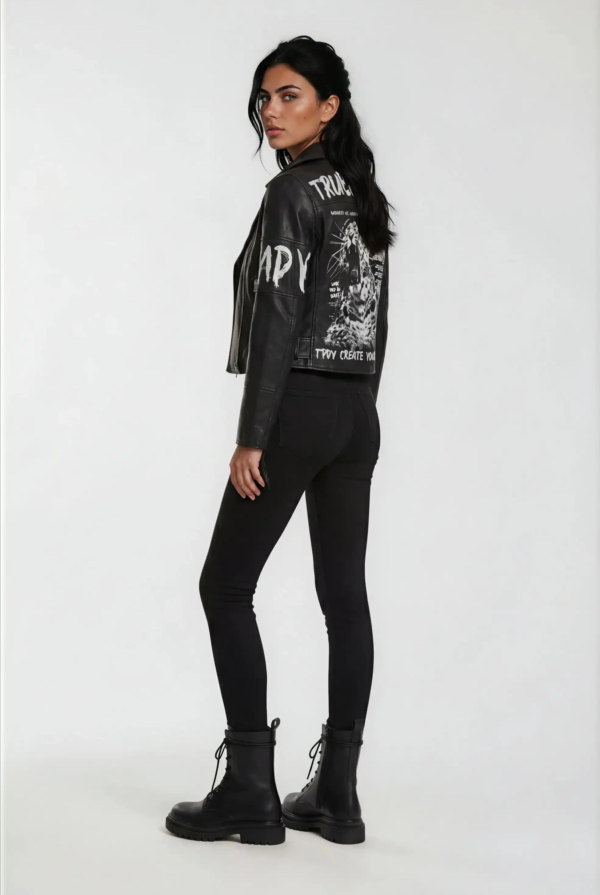 Eine Frau steht in Dreiviertel-Pose mit Blick nach links und trägt die trueprodigy Lederjacke mit Druck mit weißer Grafik auf dem Rücken, gepaart mit schwarzen Skinny Jeans und Schnürstiefeln vor einem schlichten weißen Hintergrund.
