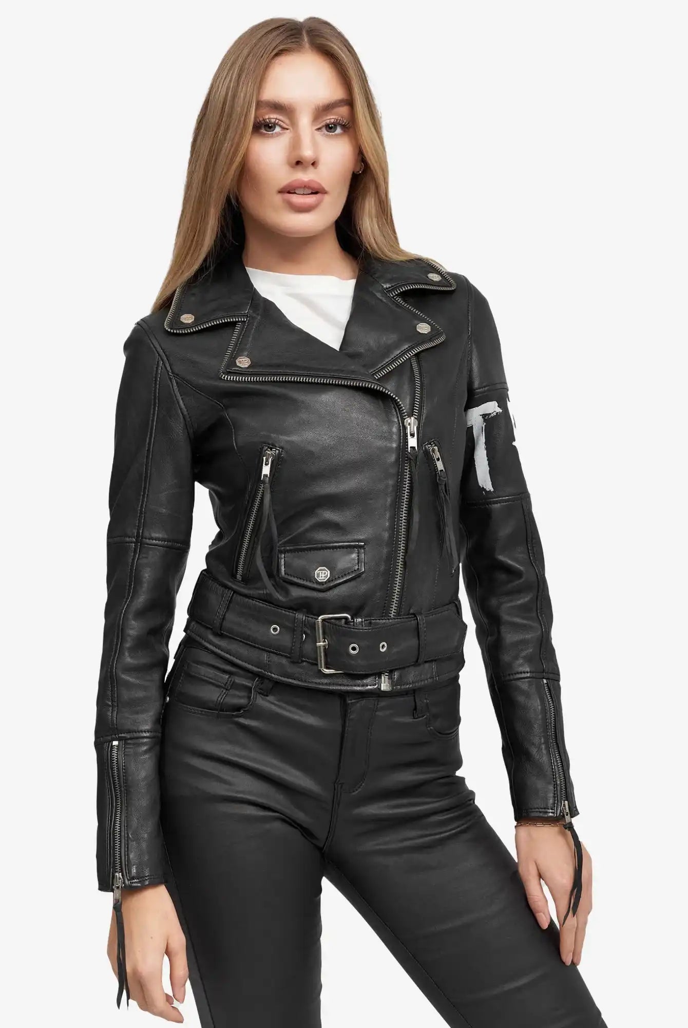 Eine Frau mit langem, glattem hellbraunem Haar trägt die Laila Lederjacke mit Druck von trueprodigy - eine schwarze Biker-Lederjacke über einem weißen Oberteil und einer schwarzen Lederhose - und steht mit dem Gesicht nach vorne vor einem schlichten weißen Hintergrund.
