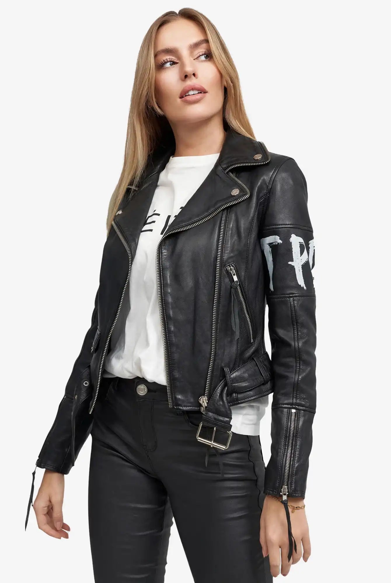 Eine Frau mit langen blonden Haaren trägt die trueprodigy Laila Lederjacke mit Druck über einem weißen Grafik-T-Shirt und einer schwarzen Hose, steht leicht links und schaut zur Seite.