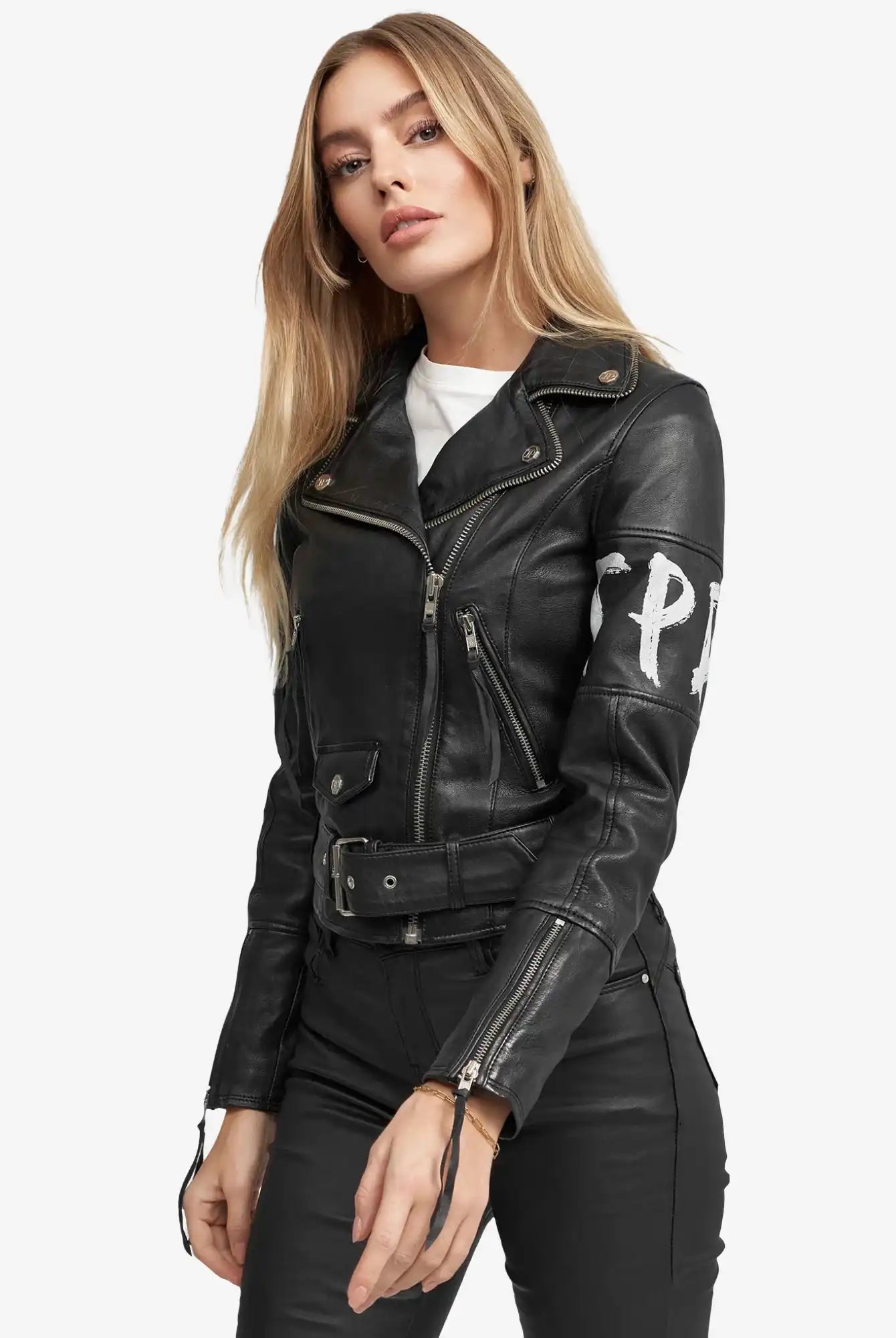 Eine Frau mit langen blonden Haaren trägt die trueprodigy Laila Lederjacke mit Druck mit Reißverschlüssen und einem Gürtel, gepaart mit einer schwarzen Hose und einem weißen Shirt. Sie steht vor einem schlichten weißen Hintergrund und schaut in die Kamera.