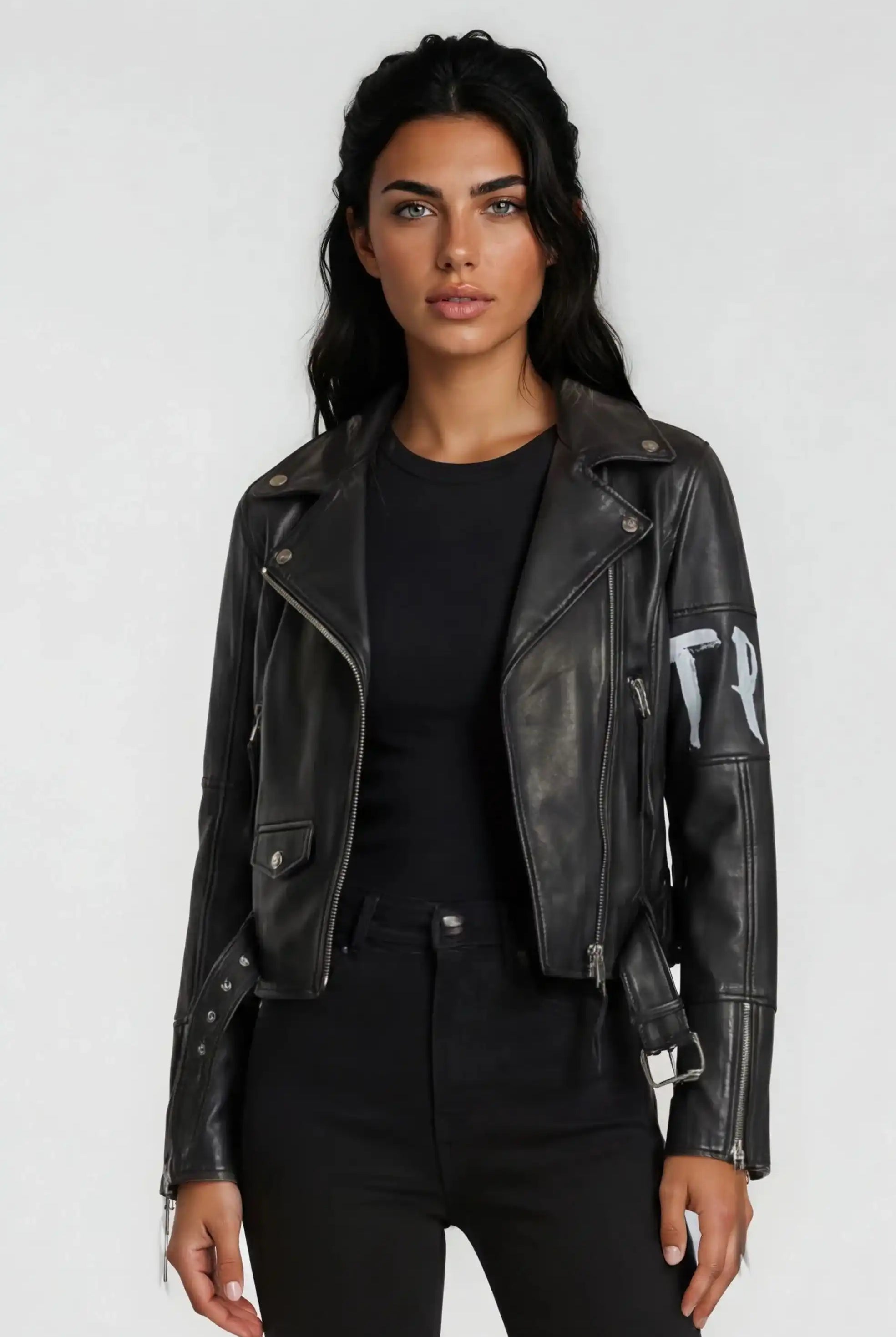 Eine Person mit langen dunklen Haaren trägt die trueprodigy Lederjacke mit Druck über einem schwarzen Hemd und einer schwarzen Hose, die vor einem schlichten hellgrauen Hintergrund steht.