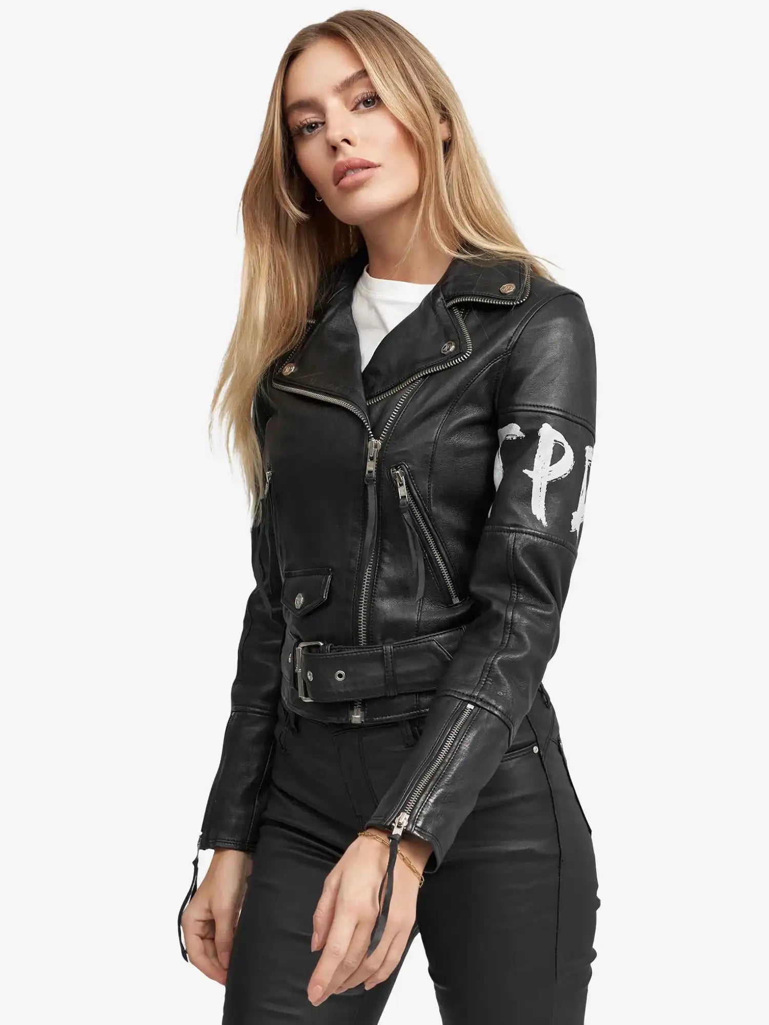 Eine Frau mit langen blonden Haaren trägt die trueprodigy Laila Lederjacke mit Druck mit Reißverschlüssen und einem Gürtel, gepaart mit einer schwarzen Hose und einem weißen Shirt. Sie steht vor einem schlichten weißen Hintergrund und schaut in die Kamera.