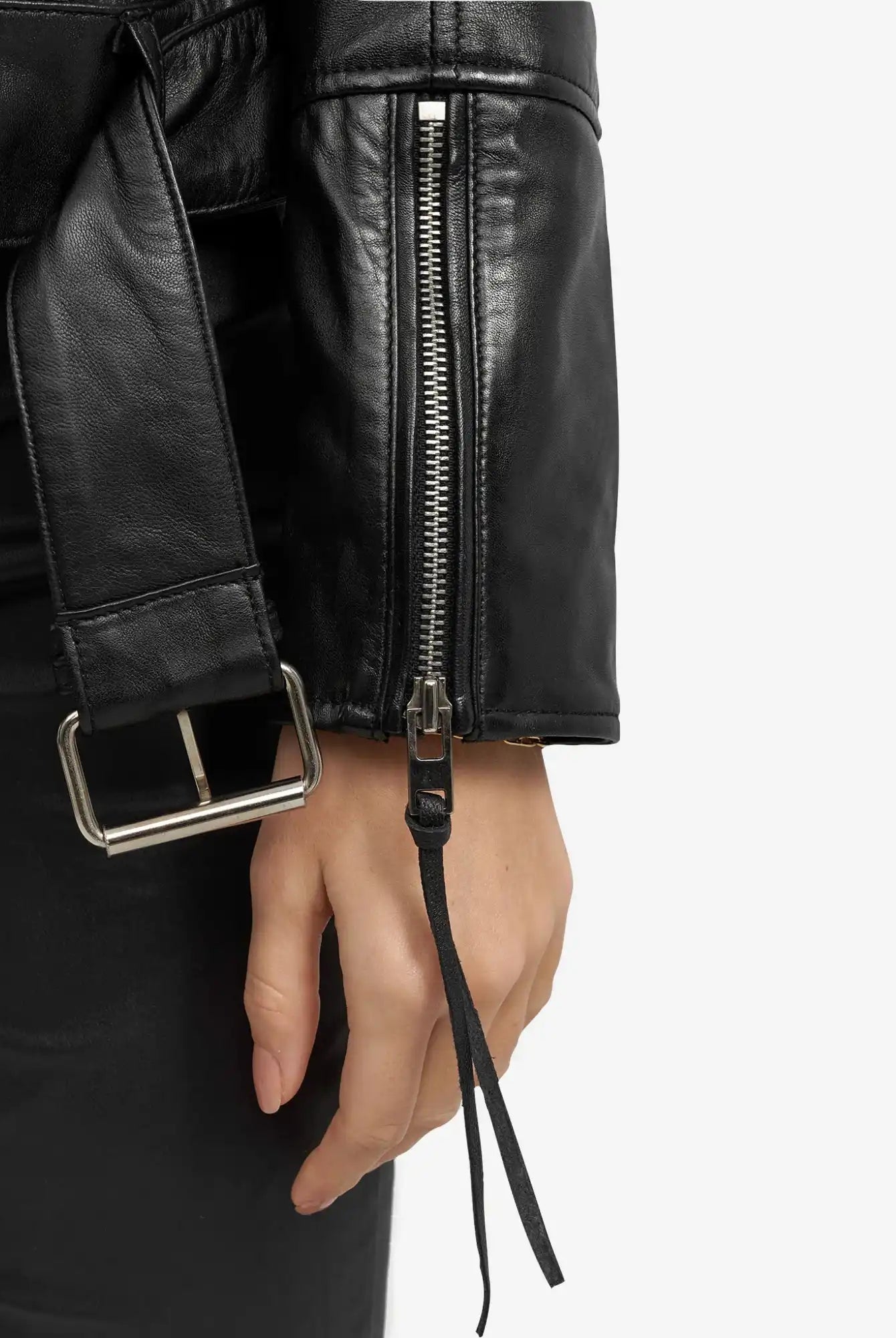 Nahaufnahme einer seitlich ruhenden Hand, die die trueprodigy Milena Lederjacke mit Gürtel trägt - schwarzes Leder mit silbernem Ärmelreißverschluss und schwarzem Gürtel mit silberner Schnalle. Der Hintergrund ist einfarbig und hell gefärbt.