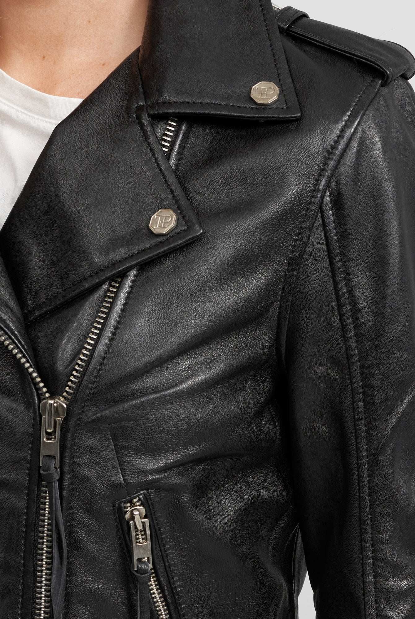 Nahaufnahme der Milena Lederjacke mit Gürtel von trueprodigy, einer schwarzen Damen-Lederjacke mit silbernen Reißverschlüssen und Druckknöpfen, gestylt über einem weißen T-Shirt. Die Aufnahme konzentriert sich auf den oberen Brust- und Schulterbereich.