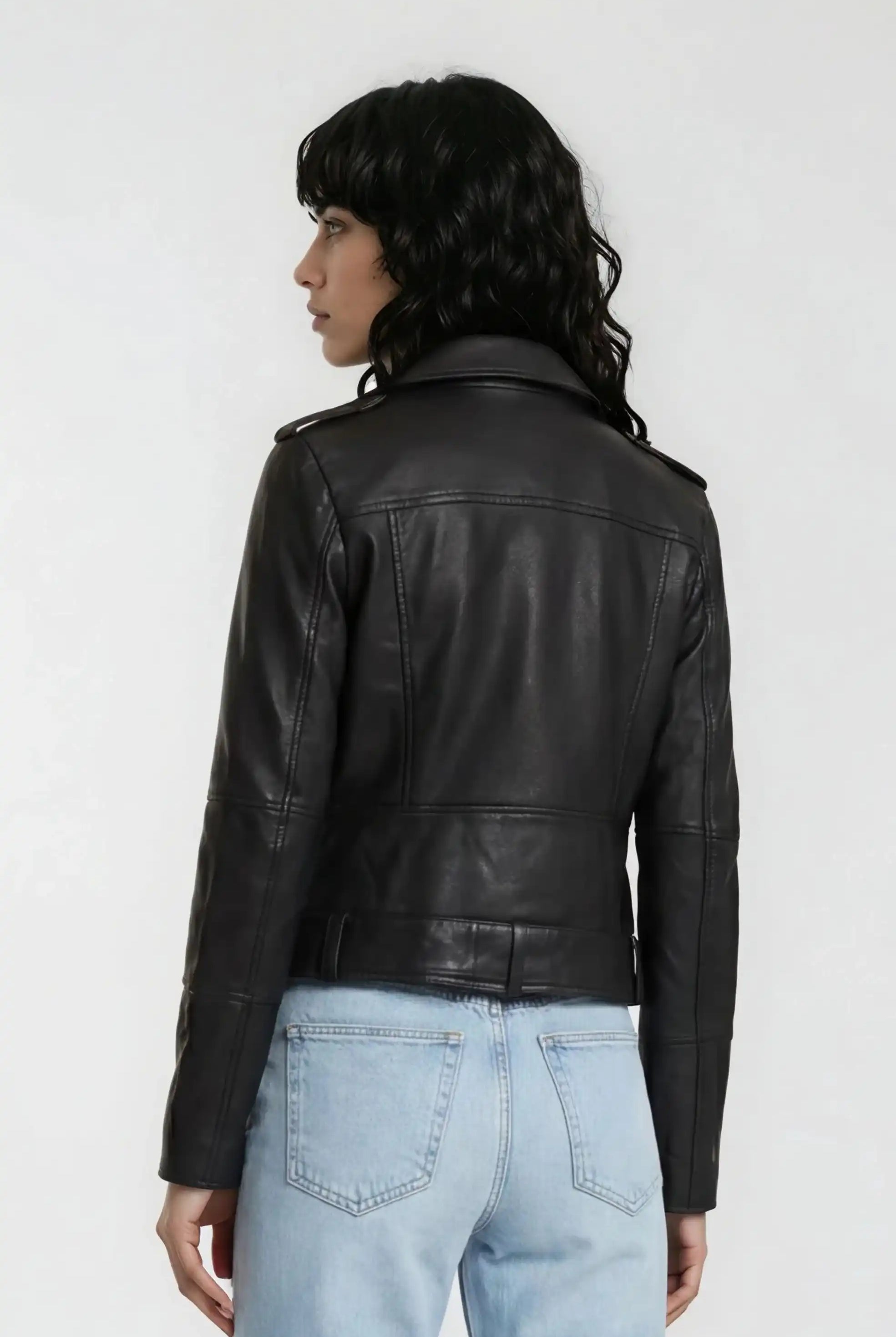 Gezeigt wird eine Person mit gewelltem dunklem Haar von hinten, die die trueprodigy Lederjacke mit Gürtel und hellblaue Jeans vor einem schlichten hellen Hintergrund trägt.