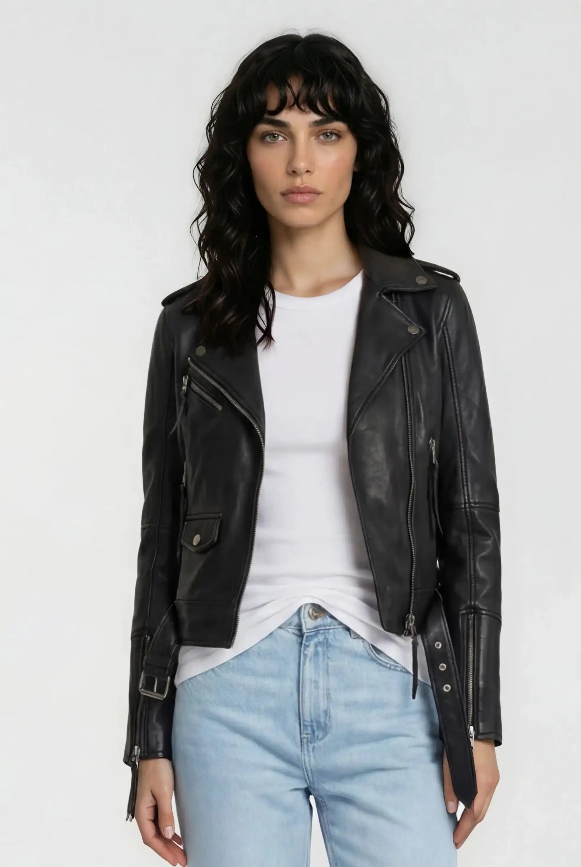 Eine Frau mit gewelltem dunklem Haar trägt die trueprodigy Lederjacke mit Gürtel über einem weißen T-Shirt und hellblauen Jeans und steht mit dem Gesicht nach vorne vor einem schlichten weißen Hintergrund.