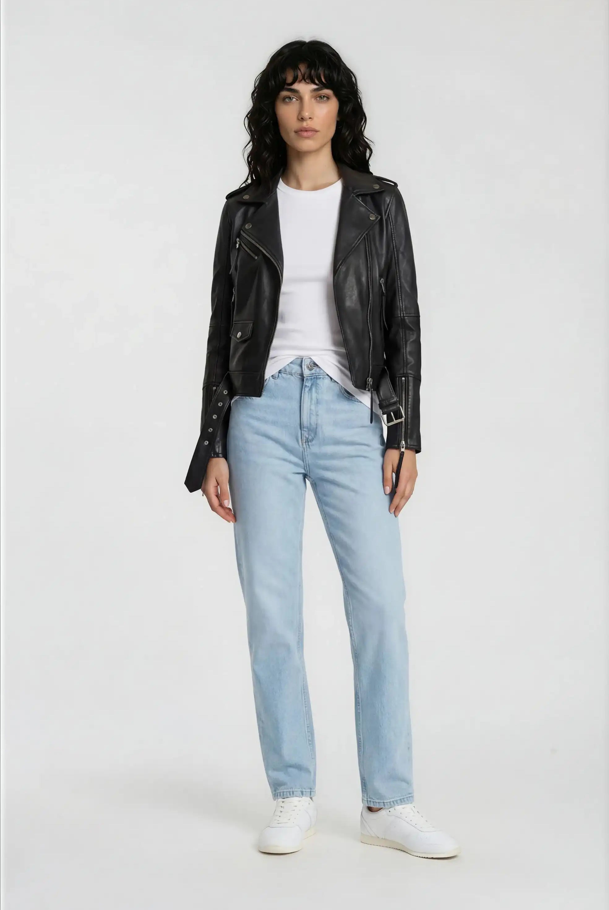 Eine Person steht mit dem Gesicht nach vorne vor einem schlichten Hintergrund und trägt die trueprodigy Lederjacke mit Gürtel über einem weißen T-Shirt, hellblaue Jeans und weiße Turnschuhe.