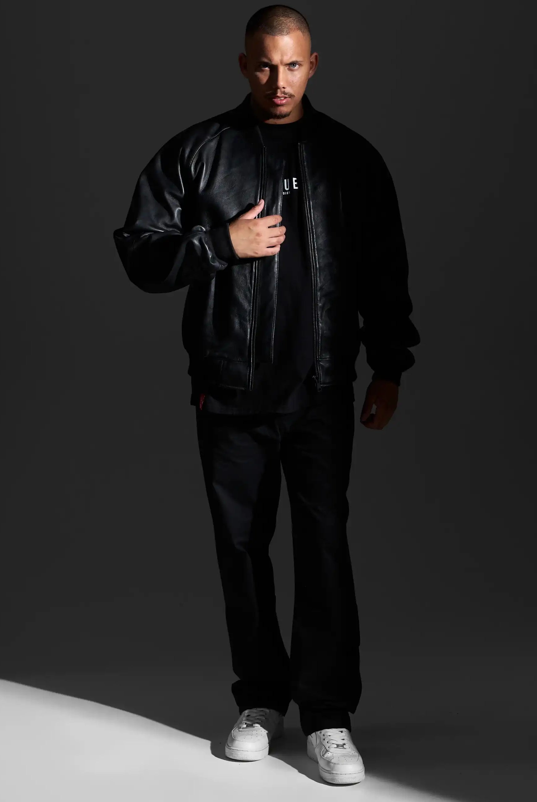 Ein Mann steht vor einem dunklen Hintergrund und trägt die trueprodigy Filip Oversized Bomber-Lederjacke, eine schwarze Hose und ein schwarzes Hemd mit weißem Schriftzug, gepaart mit weißen Sneakers. Er blickt nach vorne und hält eine Hand an den Reißverschluss seiner Jacke.