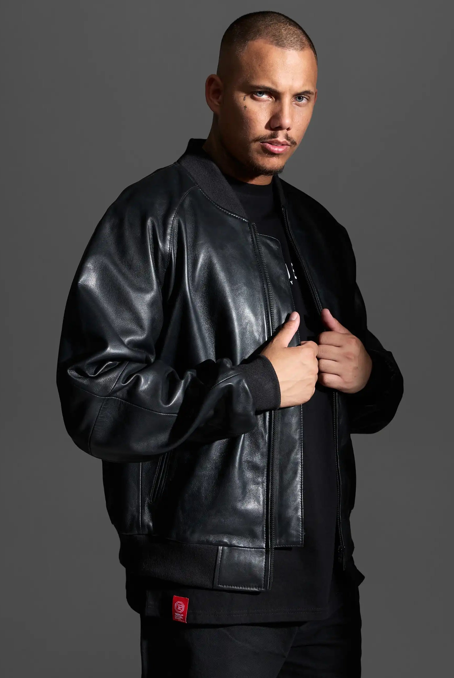 Ein Mann mit kurzen Haaren und gestutztem Schnurrbart trägt die trueprodigy Filip Oversized Bomber-Lederjacke über einem schwarzen Hemd, steht vor einem grauen Hintergrund und schaut mit neutralem Blick direkt in die Kamera.