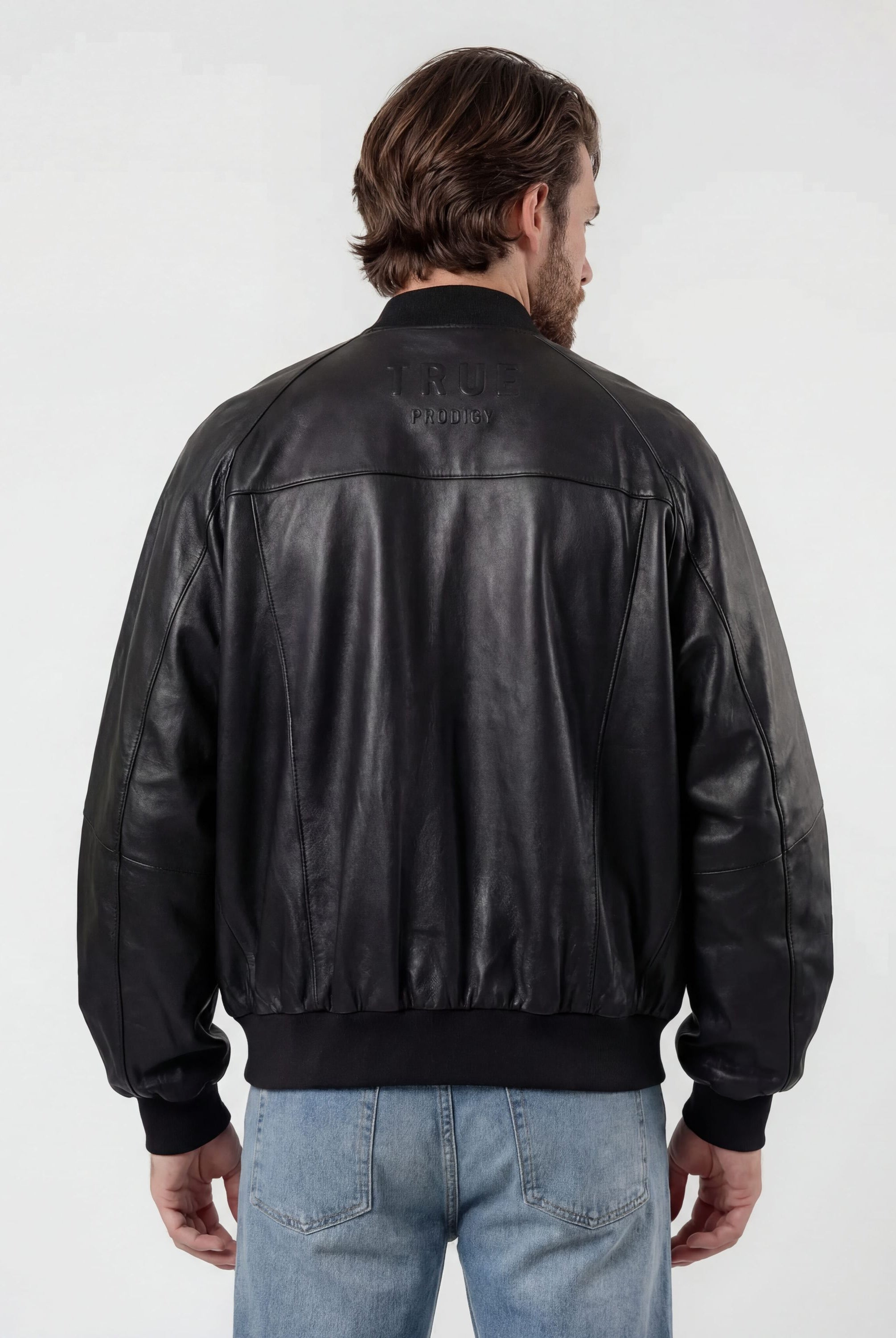 Eine Person mit hellbraunen Haaren trägt die trueprodigy Bomber Lederjacke Herren "Filip" im Oversize-Look und hellblaue Jeans und steht mit dem Rücken zur Kamera vor einem schlichten, hellen Hintergrund.