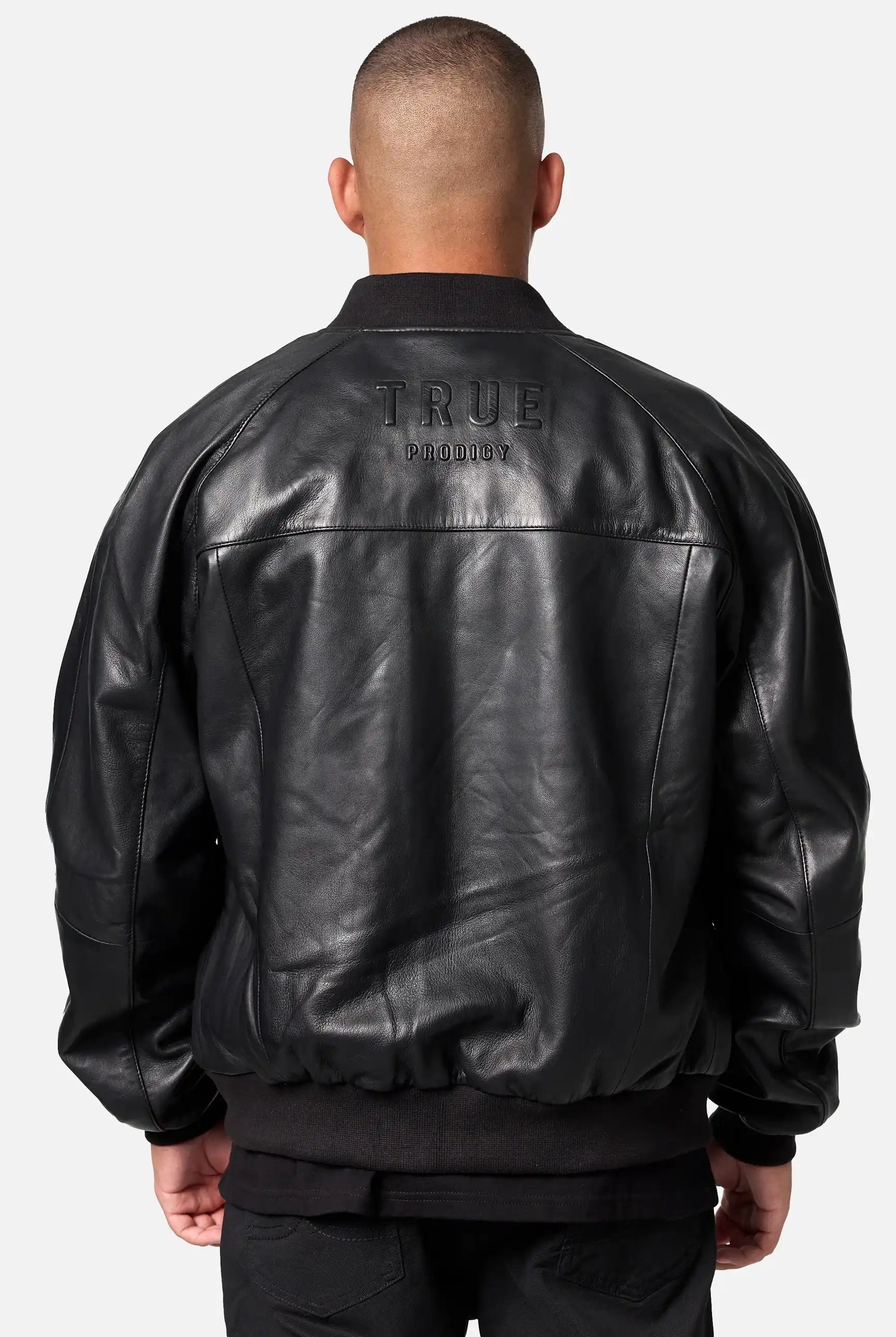 Eine Person steht mit dem Rücken zur Kamera und trägt eine trueprodigy Filip Oversized Bomber-Lederjacke mit dem großen TRUE PRODIGY-Schriftzug auf dem Rücken. Der Hintergrund ist hell und schlicht.
