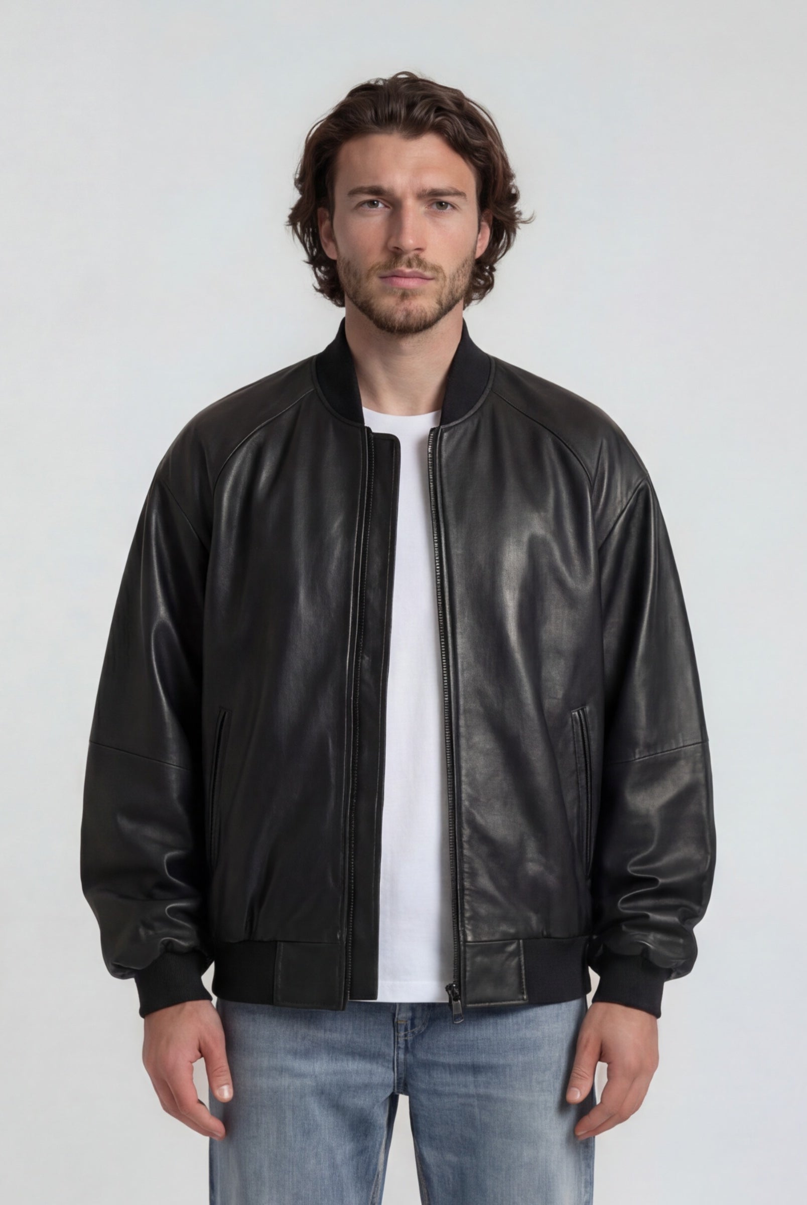 Ein Mann mit gewellten braunen Haaren und kurzem Bart steht vor einem schlichten Hintergrund und trägt die trueprodigy Bomber Lederjacke Herren "Filip" im Oversize-Look aus Schafsleder, gepaart mit einem weißen T-Shirt und hellblauen Jeans.