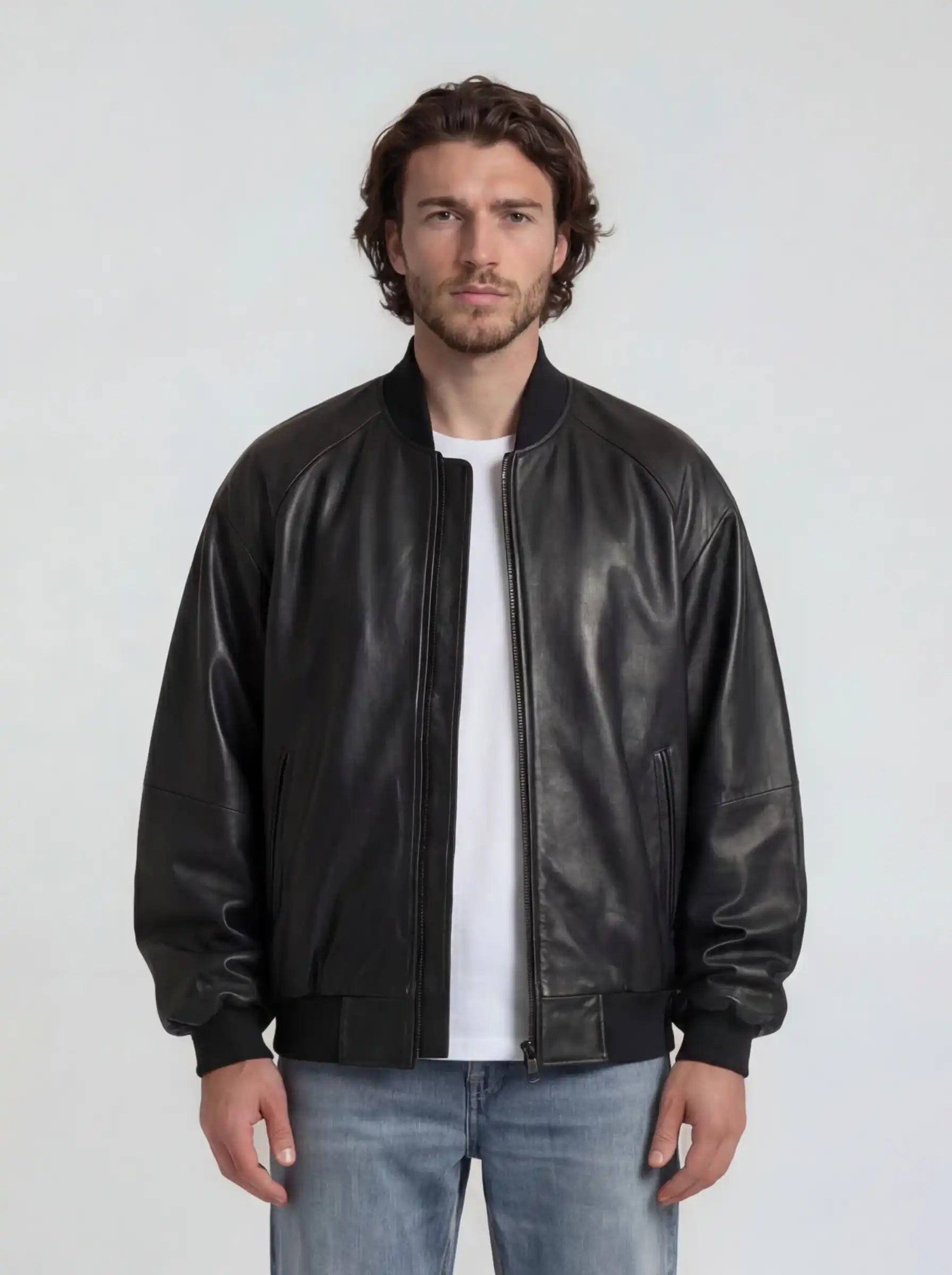 Ein Mann mit gewellten braunen Haaren und kurzem Bart steht vor einem schlichten Hintergrund und trägt die trueprodigy Bomber Lederjacke Herren "Filip" im Oversize-Look aus Schafsleder, gepaart mit einem weißen T-Shirt und hellblauen Jeans.