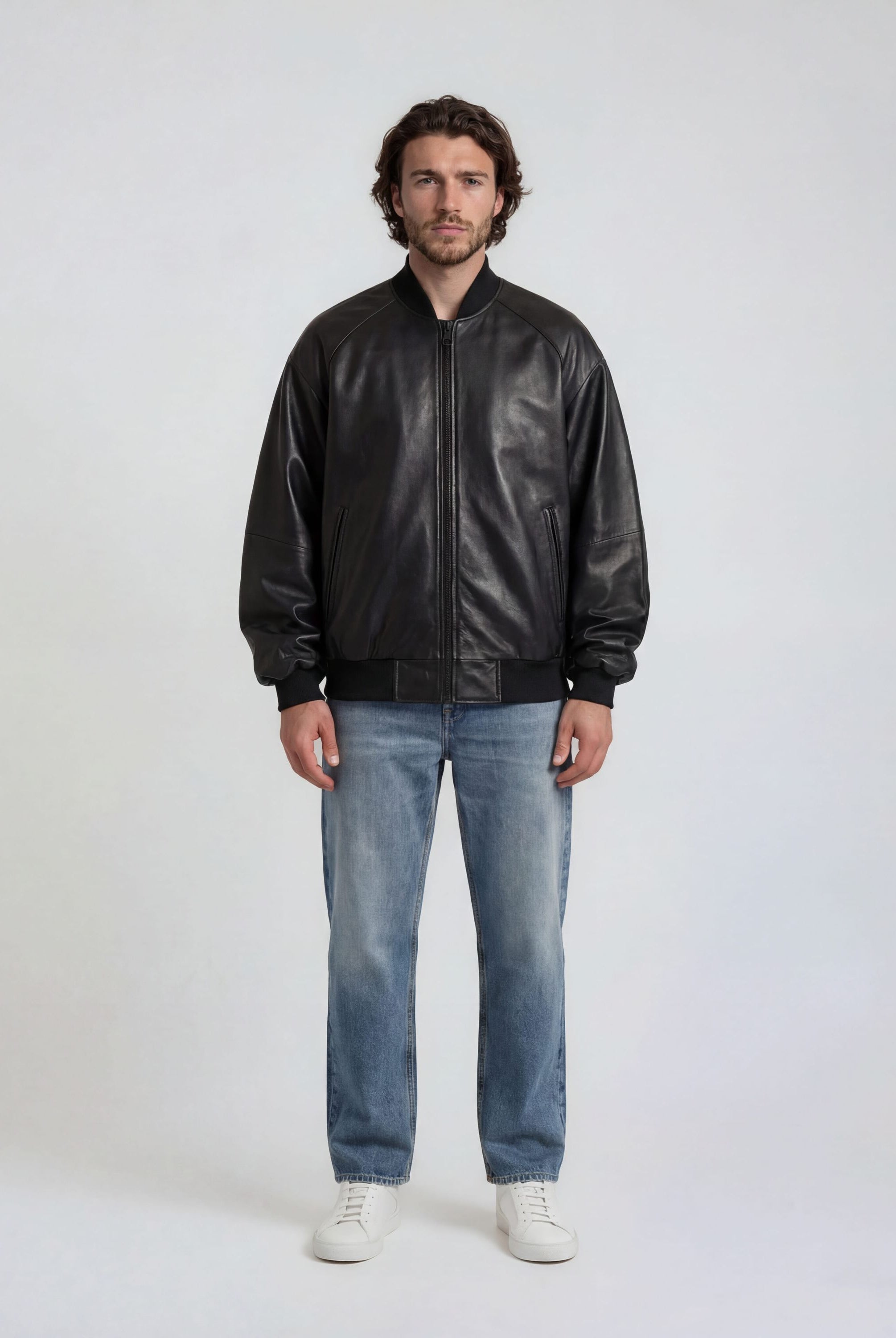 Ein Mann steht vor einem schlichten, hellen Hintergrund. Er trägt die trueprodigy Bomber Lederjacke Herren "Filip" im Oversize-Look, blaue Jeans und weiße Sneakers.