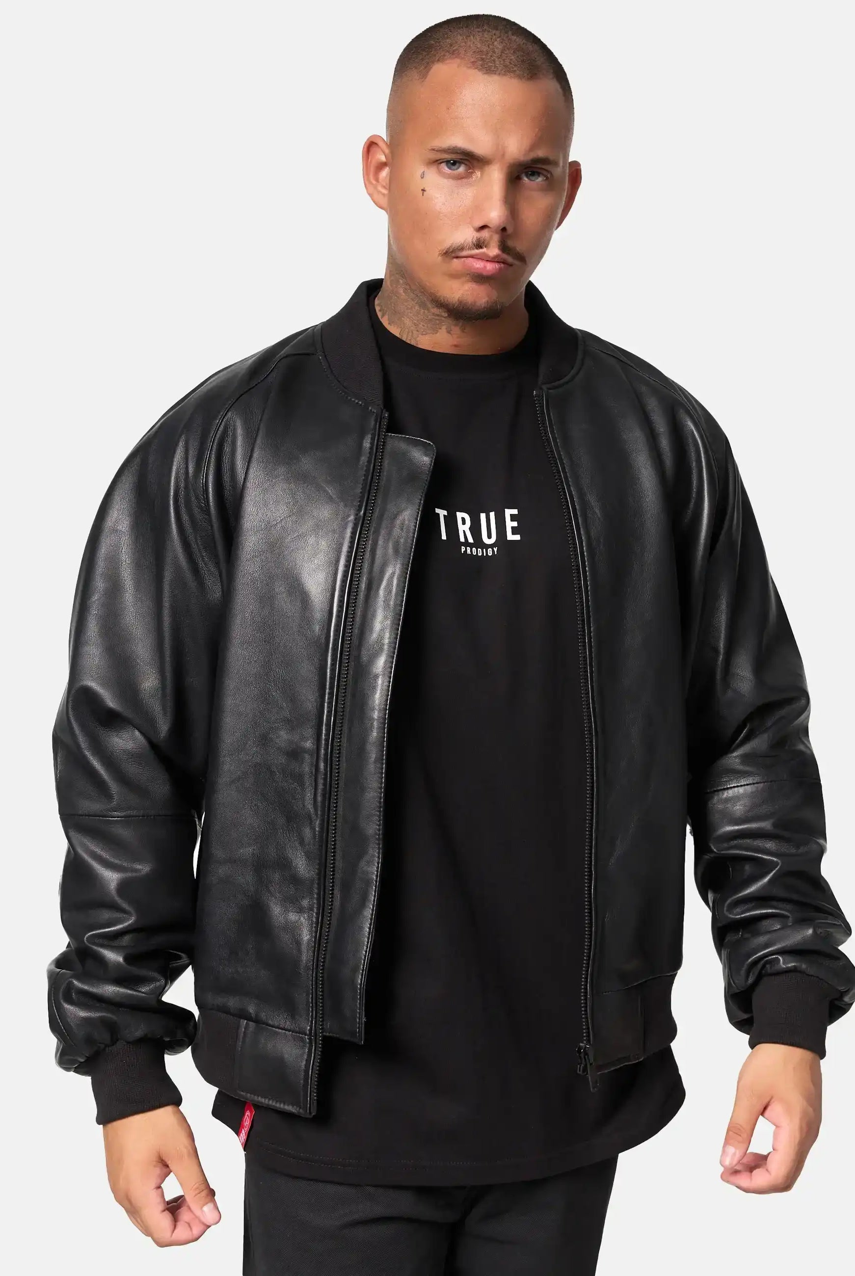 Ein Mann mit kurzen Haaren und gestutztem Bart trägt eine schwarze trueprodigy Filip Oversized Bomber-Lederjacke über einem schwarzen Hemd mit dem Aufdruck "TRUE", stehend vor einem schlichten hellen Hintergrund.