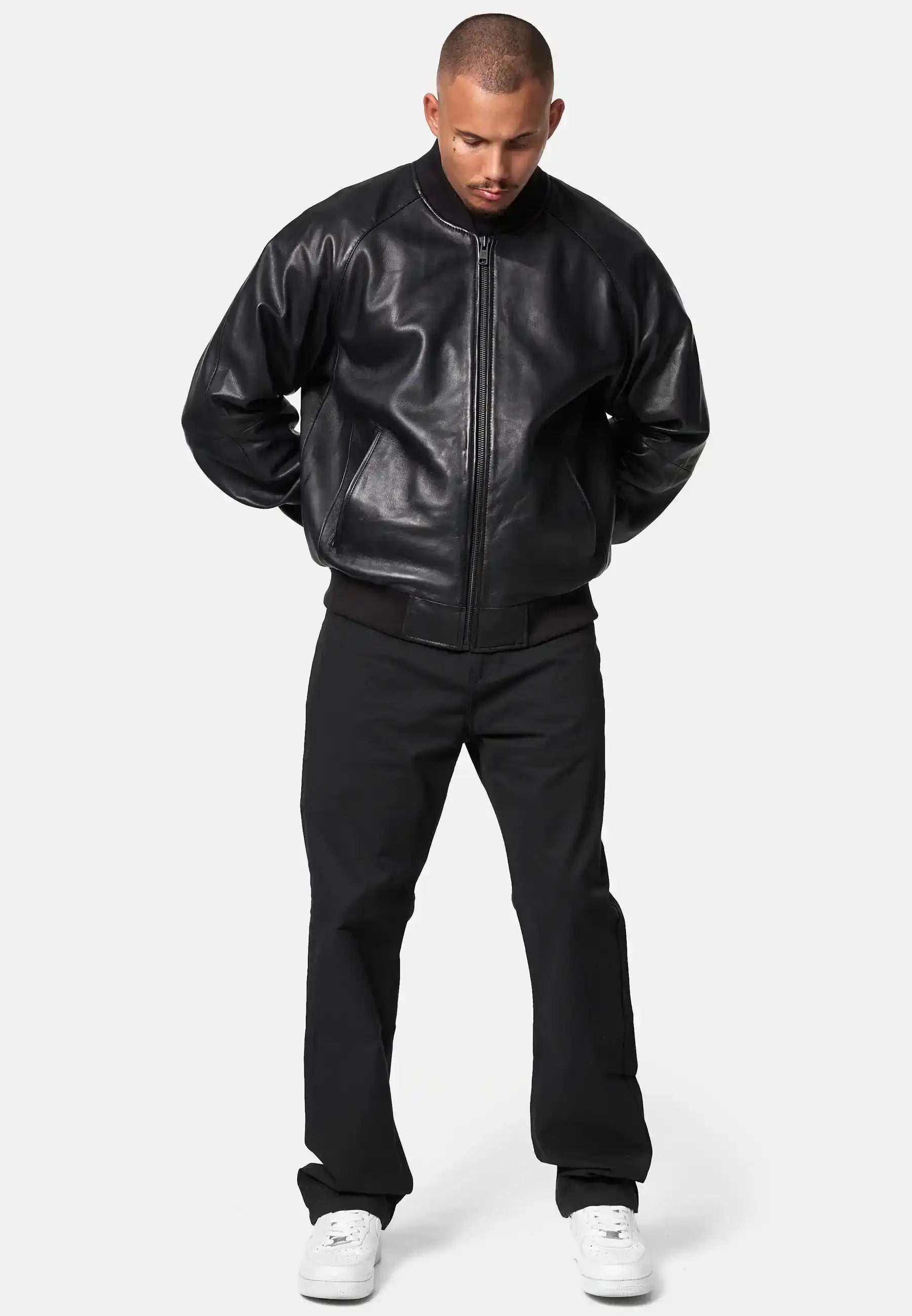 Ein Mann blickt mit leicht gesenktem Kopf nach vorne, die Hände auf dem Rücken, und trägt die trueprodigy Filip Oversized Bomber-Lederjacke aus schwarzem Leder, eine schwarze Hose und weiße Turnschuhe vor einem schlichten hellen Hintergrund.
