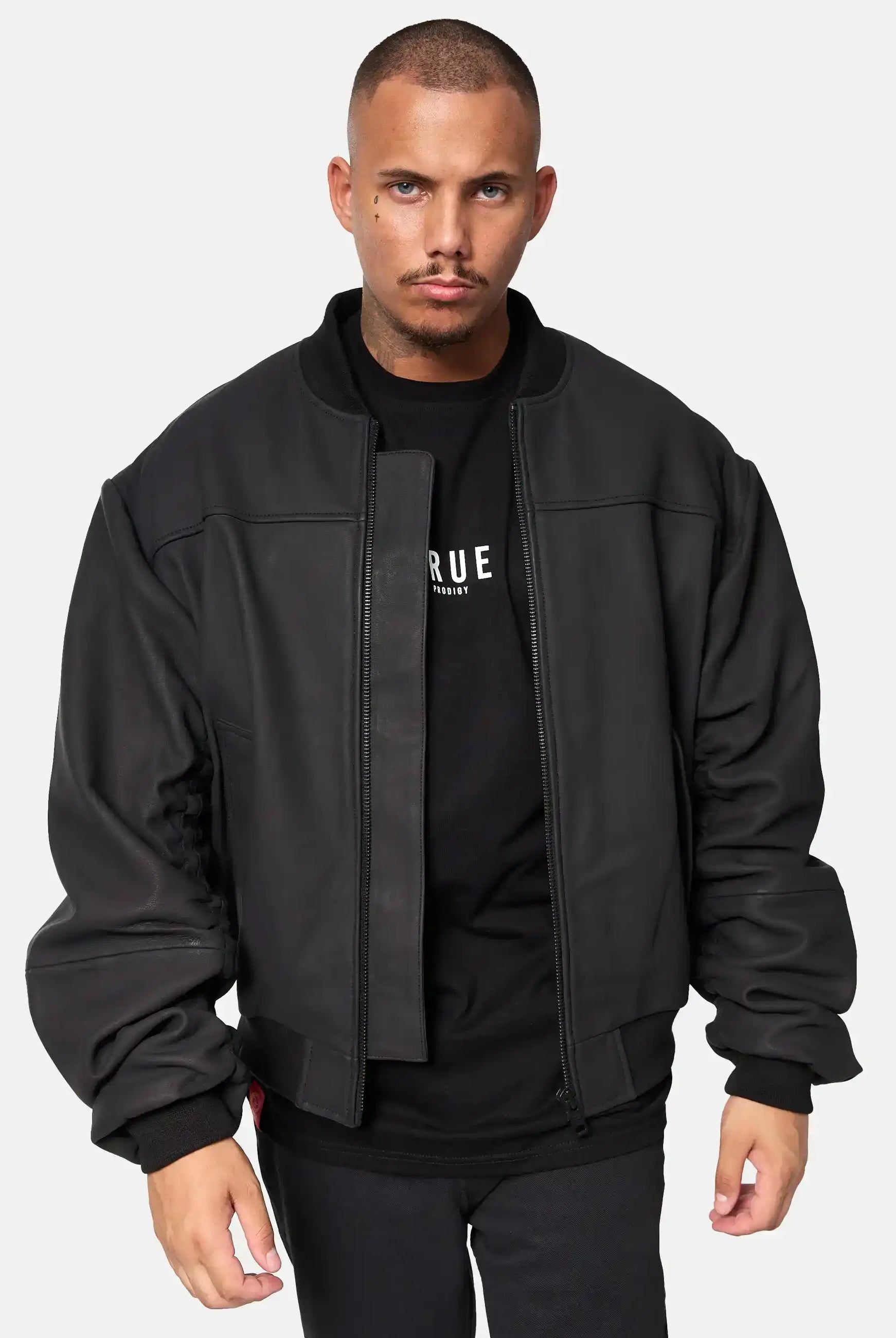 Ein Mann mit Kurzhaarschnitt und Schnurrbart trägt eine schwarze Nazar Oversized Bomber-Lederjacke matt von trueprodigy über einem schwarzen trueprodigy-Shirt mit weißem Text, steht vor einem schlichten weißen Hintergrund und blickt mit neutralem Ausdruck nach vorne.