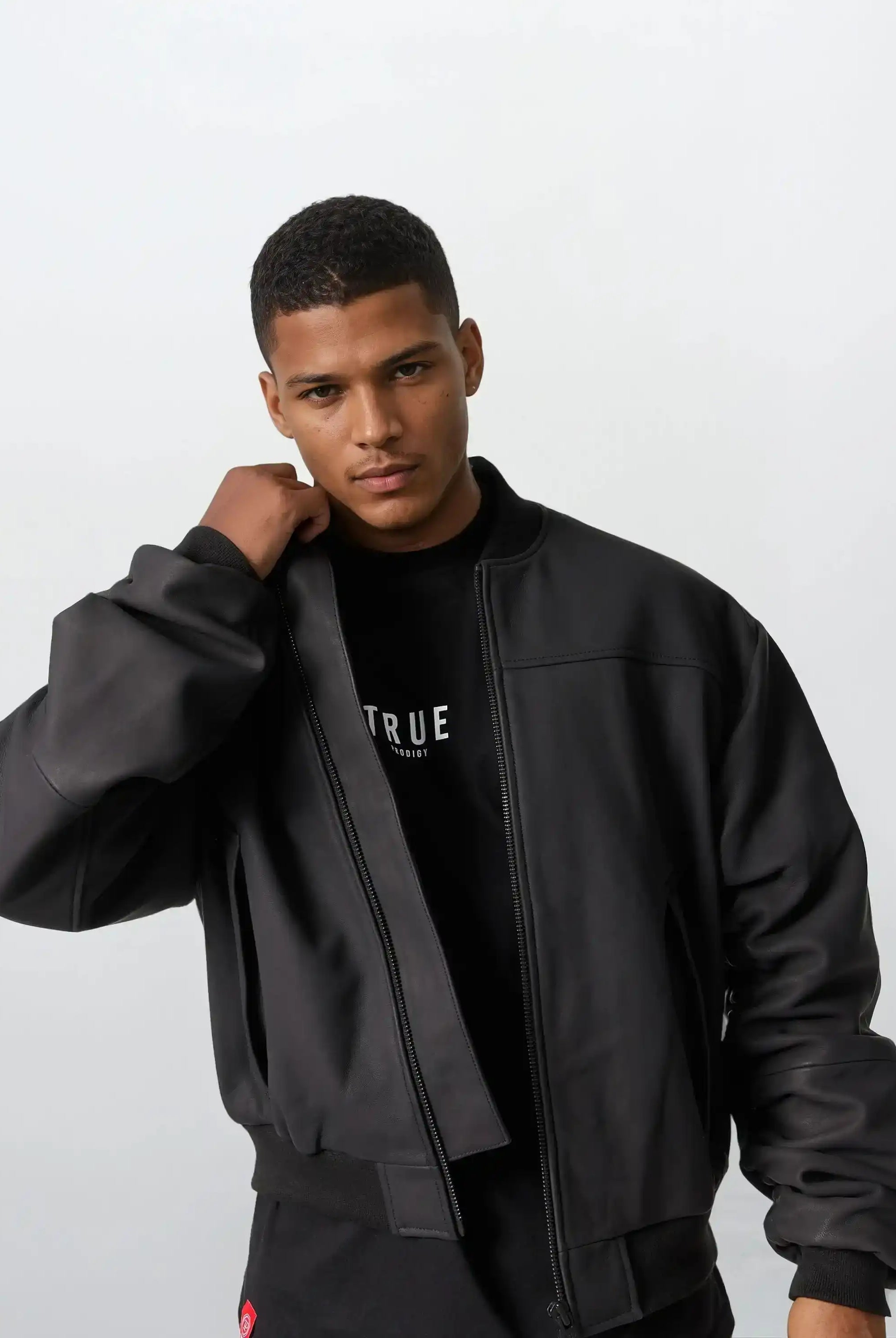 Ein Mann mit kurzen Haaren trägt die Nazar Oversized Bomber-Lederjacke matt von trueprodigy über einem schwarzen T-Shirt mit weißem Text, blickt in die Kamera und richtet seine Jacke vor hellem Hintergrund.