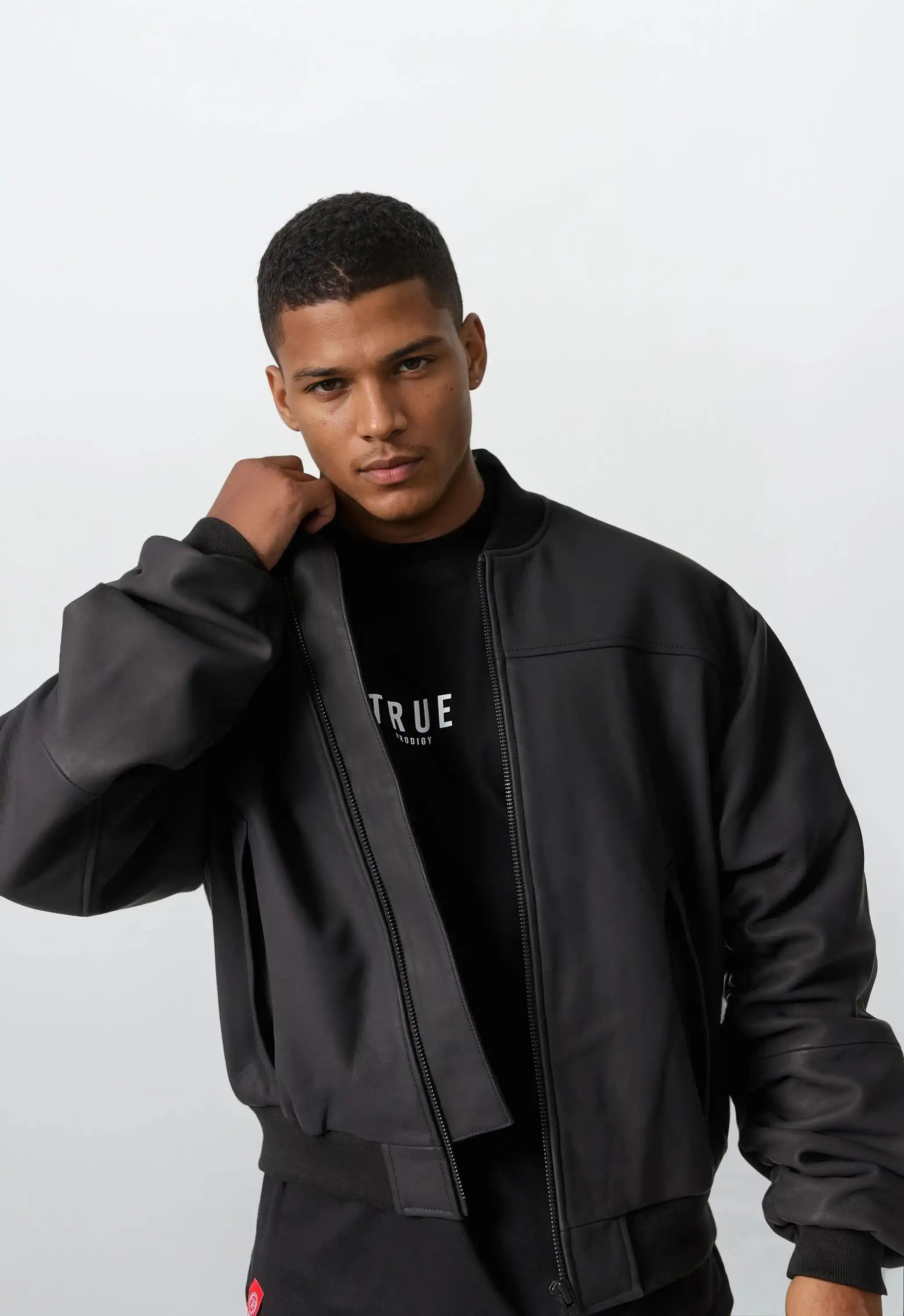 Ein Mann mit kurzen Haaren trägt die Nazar Oversized Bomber-Lederjacke matt von trueprodigy über einem schwarzen T-Shirt mit weißem Text, blickt in die Kamera und richtet seine Jacke vor hellem Hintergrund.