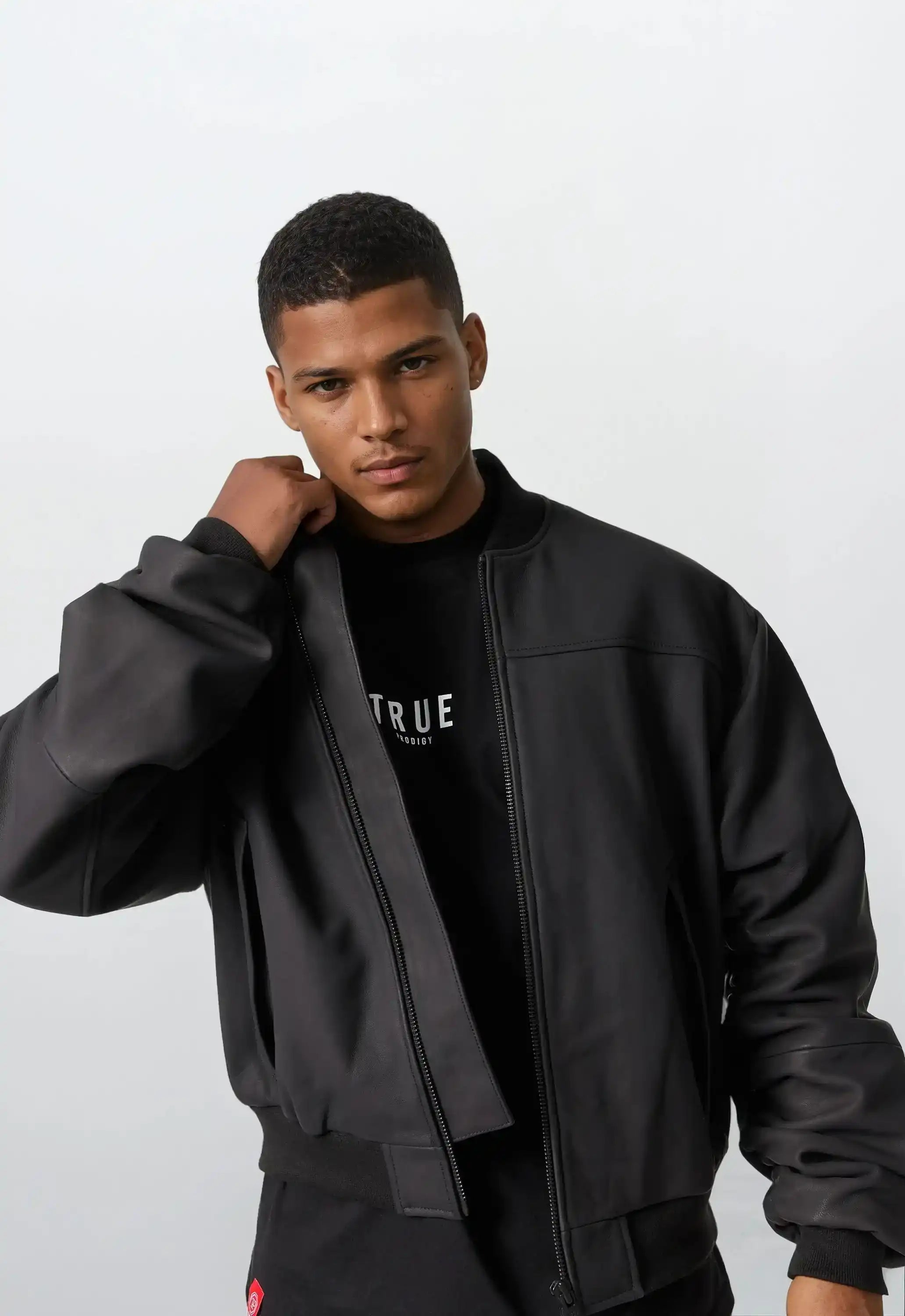 Ein Mann mit kurzen Haaren trägt die Nazar Oversized Bomber-Lederjacke matt von trueprodigy über einem schwarzen T-Shirt mit weißem Text, blickt in die Kamera und richtet seine Jacke vor hellem Hintergrund.