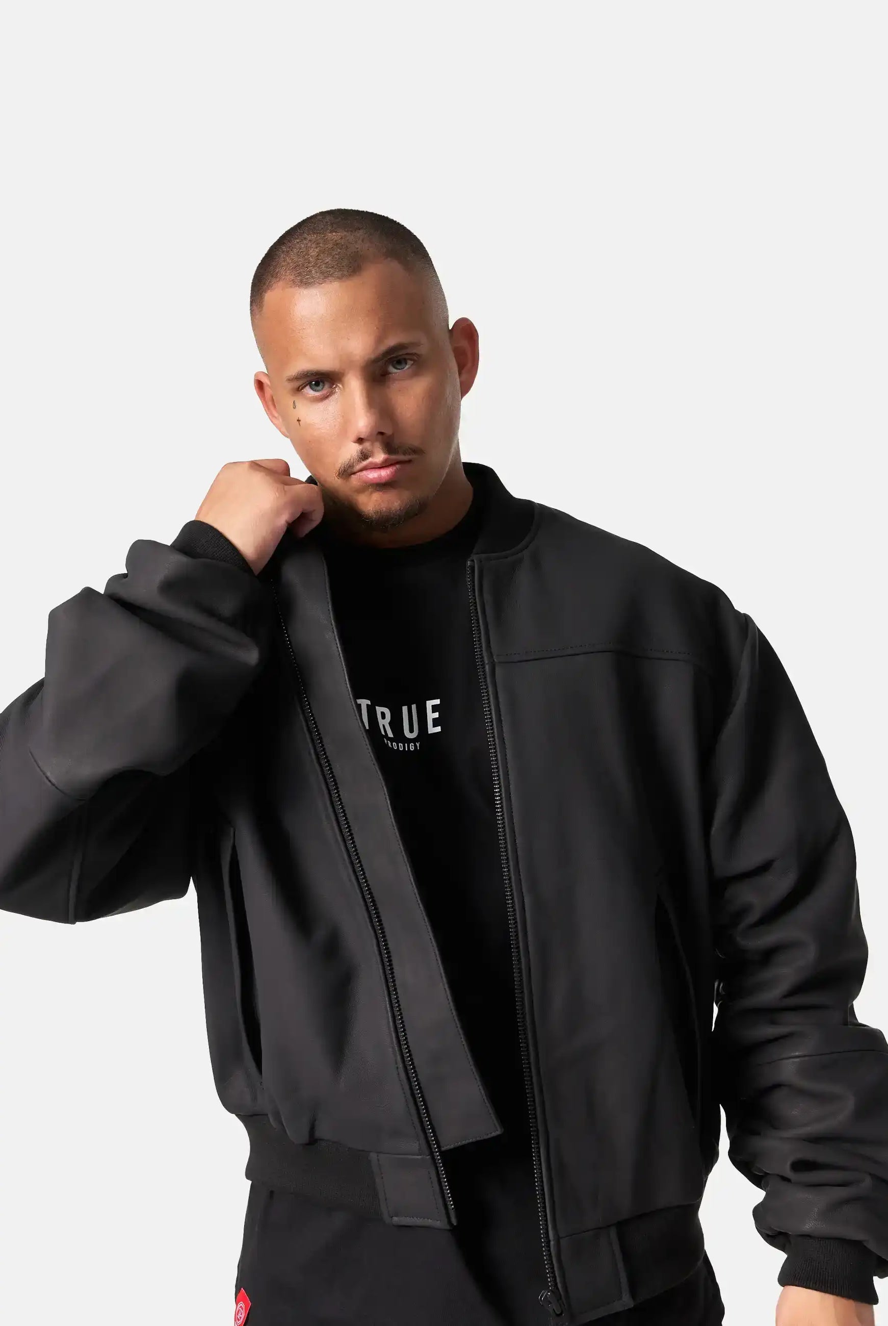 Ein Mann mit kurzem Haar und getrimmtem Bart trägt die Nazar Oversized Bomber-Lederjacke matt von trueprodigy offen über einem schwarzen Shirt mit weißer Schrift und blickt direkt in die Kamera. Der Hintergrund ist schlicht weiß.