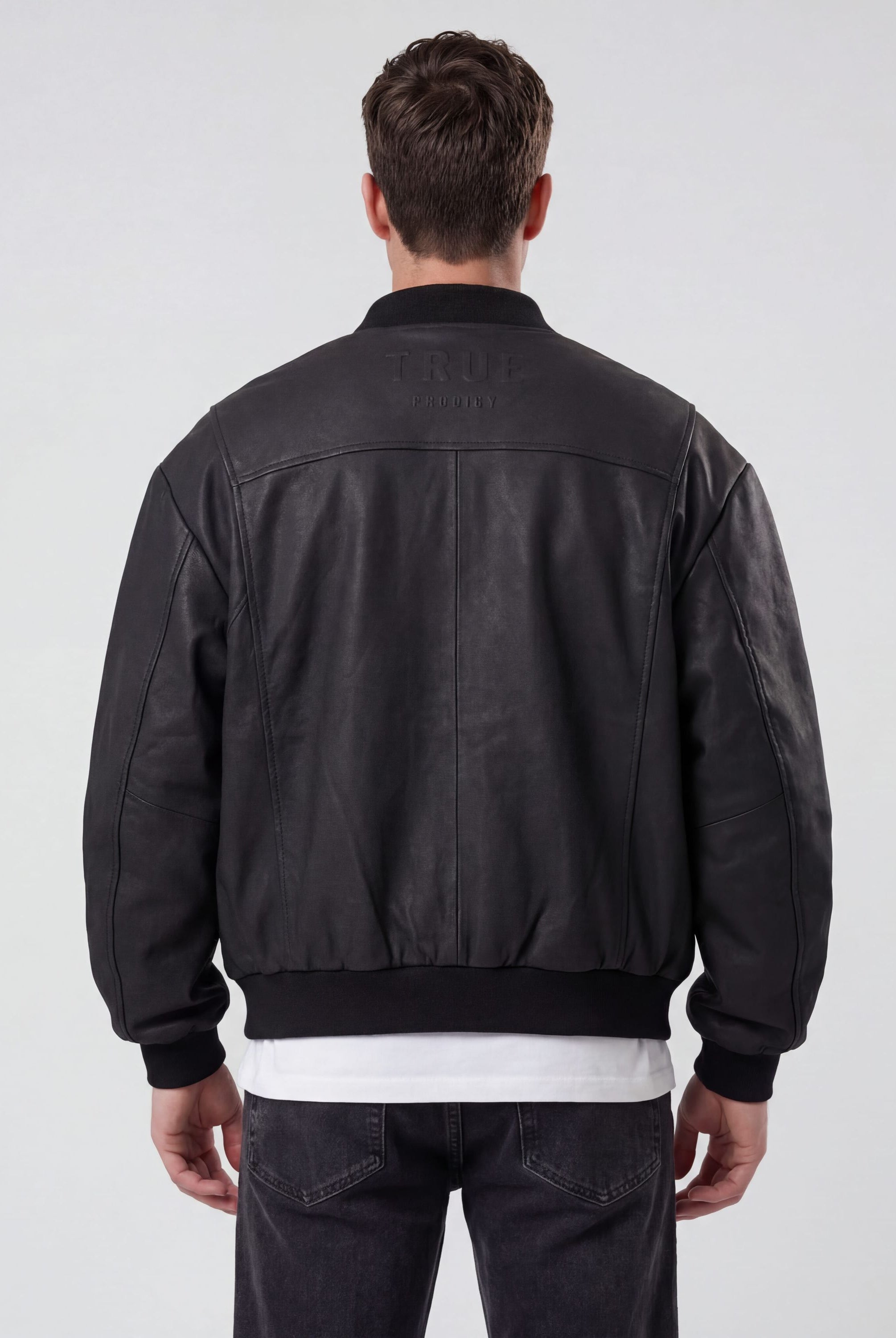 Ein Mann steht mit dem Rücken zur Kamera und trägt die trueprodigy Bomber Lederjacke Herren "Nazar" im Oversize-Schnitt in schwarz, über einem weißen Hemd und einer dunklen Jeans. Der Hintergrund ist einfarbig und hell gefärbt.