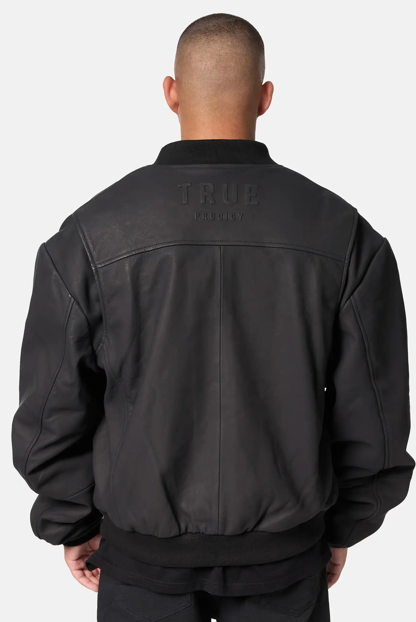 Eine Person mit kurzen Haaren, von hinten gesehen, trägt eine schwarze Nazar Oversized Bomber-Lederjacke matt von trueprodigy. Auf dem oberen Rücken ist "trueprodigy" vor einem schlichten hellen Hintergrund eingeprägt.