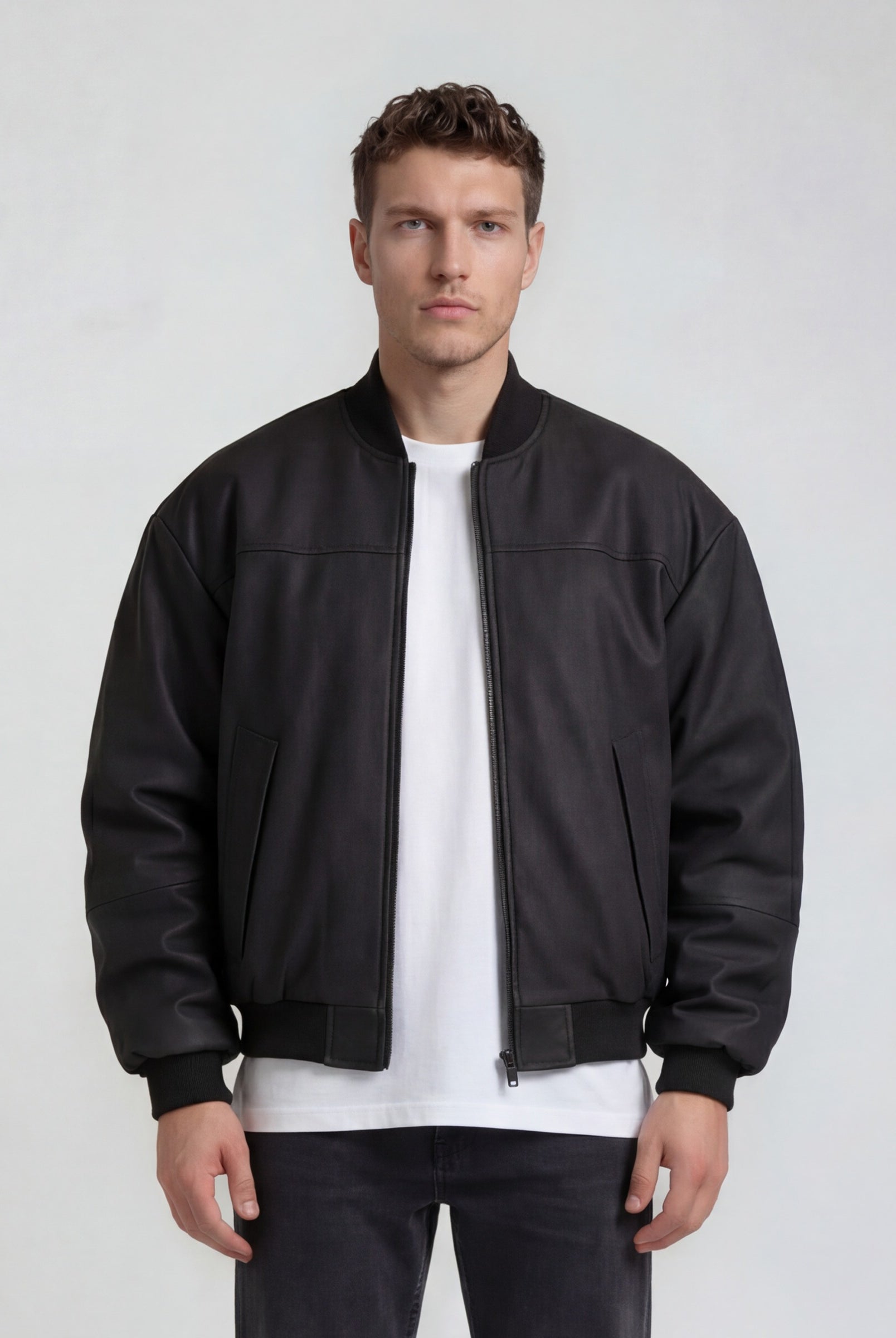 Ein Mann sieht nach vorne und trägt die trueprodigy Bomber Lederjacke Herren "Nazar" im Oversize-Schnitt in Schwarz über einem weißen T-Shirt und schwarzen Jeans vor hellem Hintergrund.