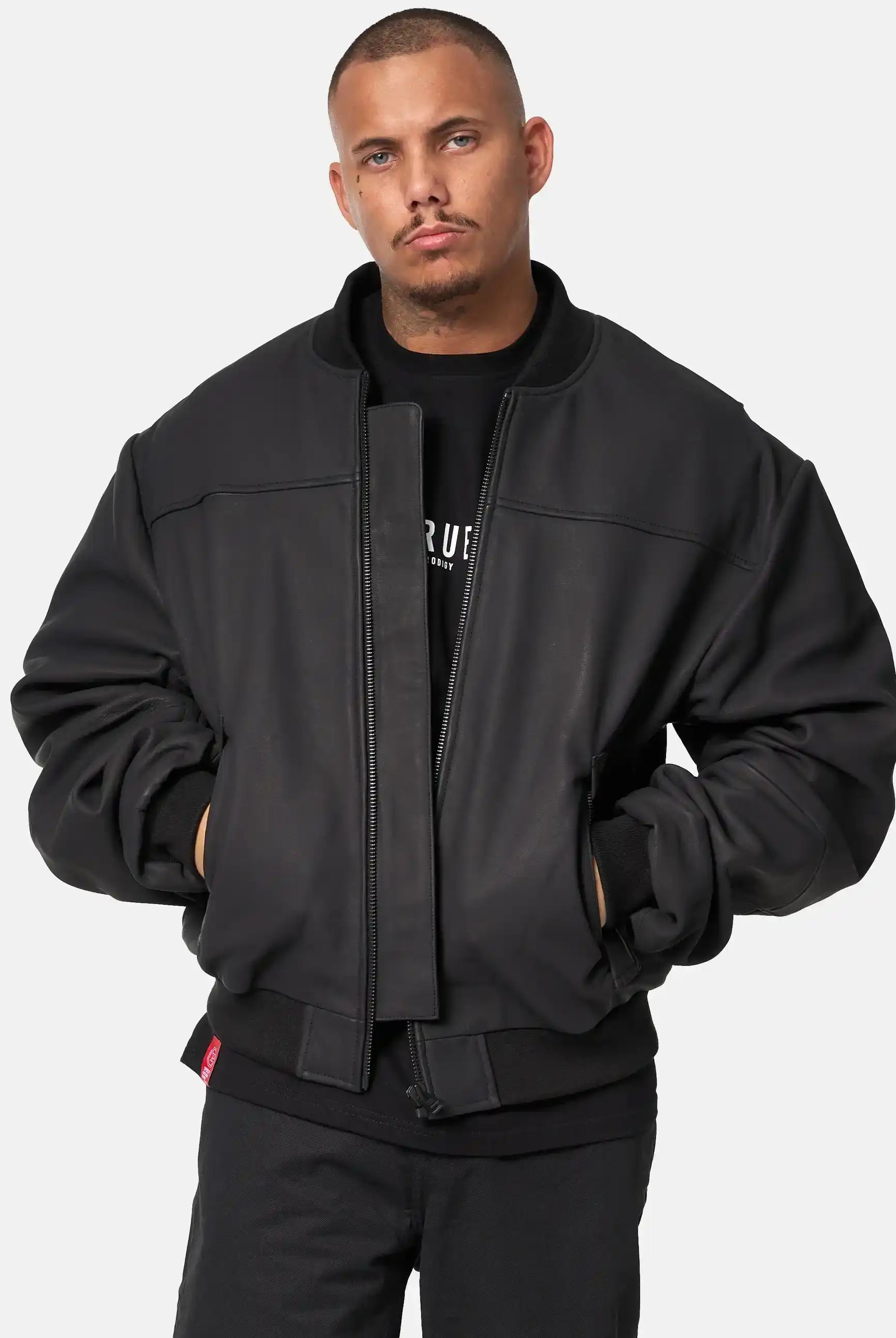 Eine Person mit kurzen Haaren und gestutzter Gesichtsbehaarung steht vor einem schlichten Hintergrund, Hände in den Taschen, trägt eine schwarze Nazar Oversized Bomber-Lederjacke matt von trueprodigy über einem schwarzen Hemd und einer schwarzen Hose und hat einen neutralen Gesichtsausdruck.