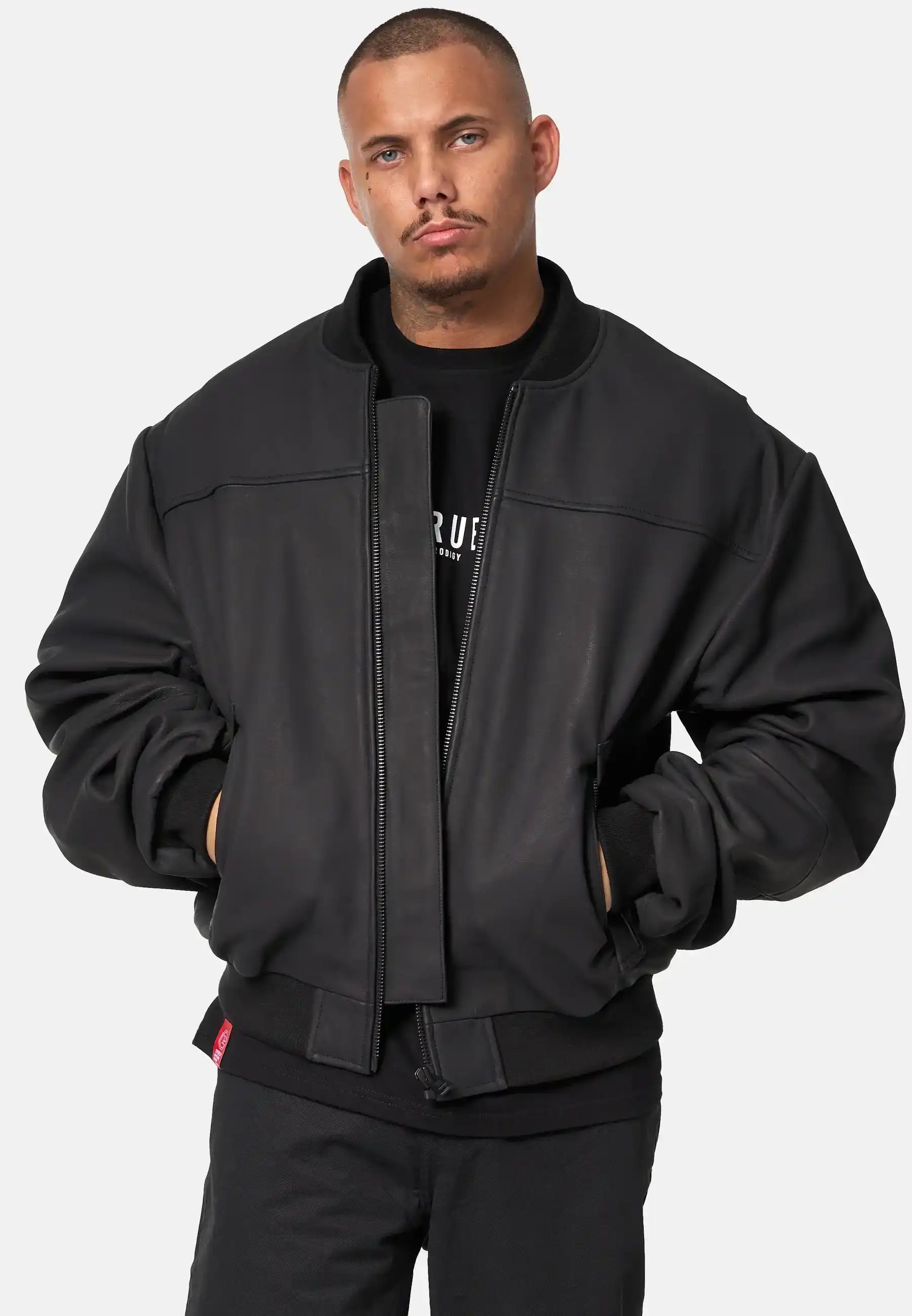 Eine Person mit kurzen Haaren und gestutzter Gesichtsbehaarung steht vor einem schlichten Hintergrund, Hände in den Taschen, trägt eine schwarze Nazar Oversized Bomber-Lederjacke matt von trueprodigy über einem schwarzen Hemd und einer schwarzen Hose und hat einen neutralen Gesichtsausdruck.