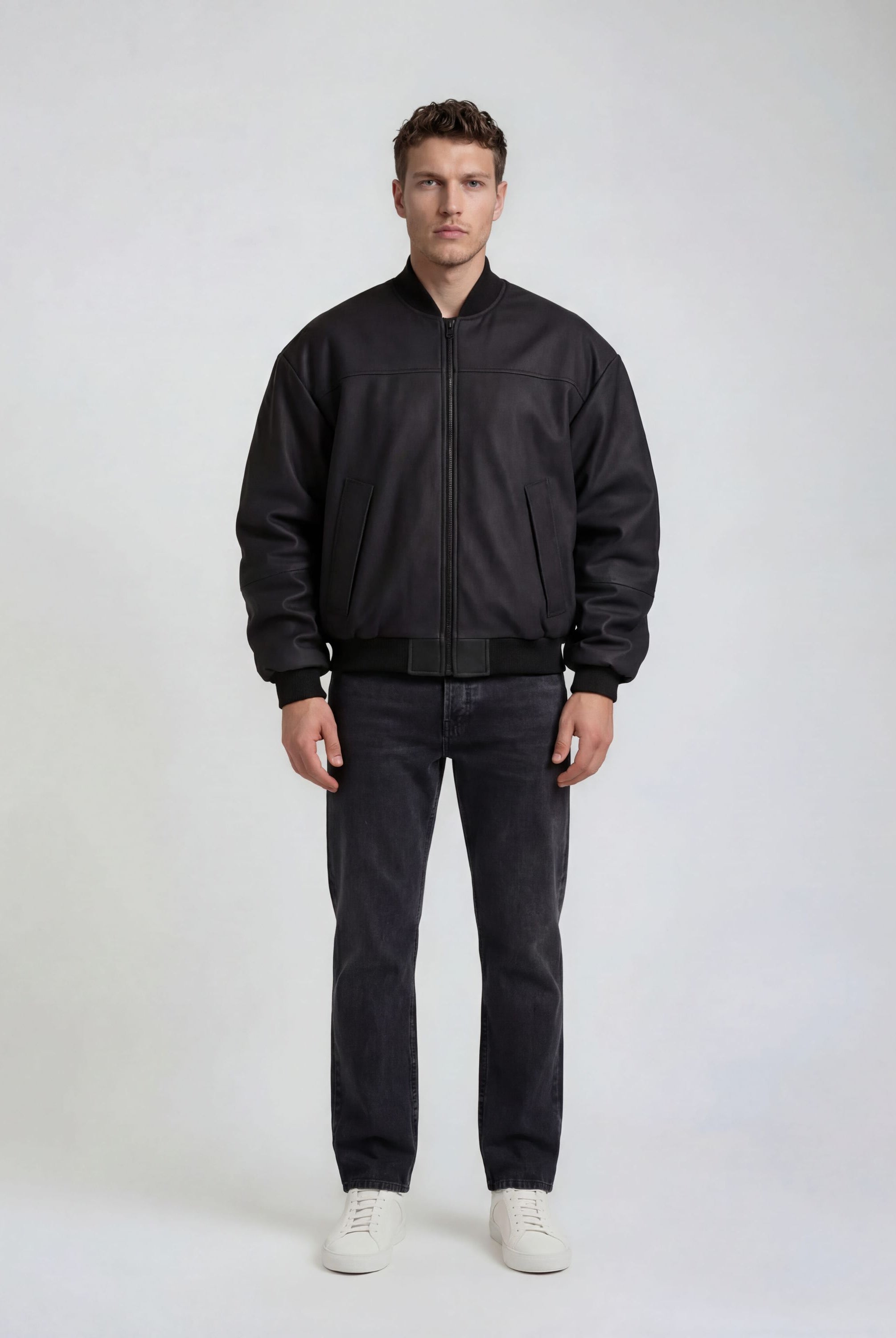 Ein Mann steht vor einem schlichten, hellen Hintergrund und trägt die trueprodigy Bomber Lederjacke Herren "Nazar" im Oversize-Schnitt, gepaart mit einer dunklen Jeans und weißen Sneakers. Seine Arme hängen entspannt an seinen Seiten und sein Gesichtsausdruck ist neutral.