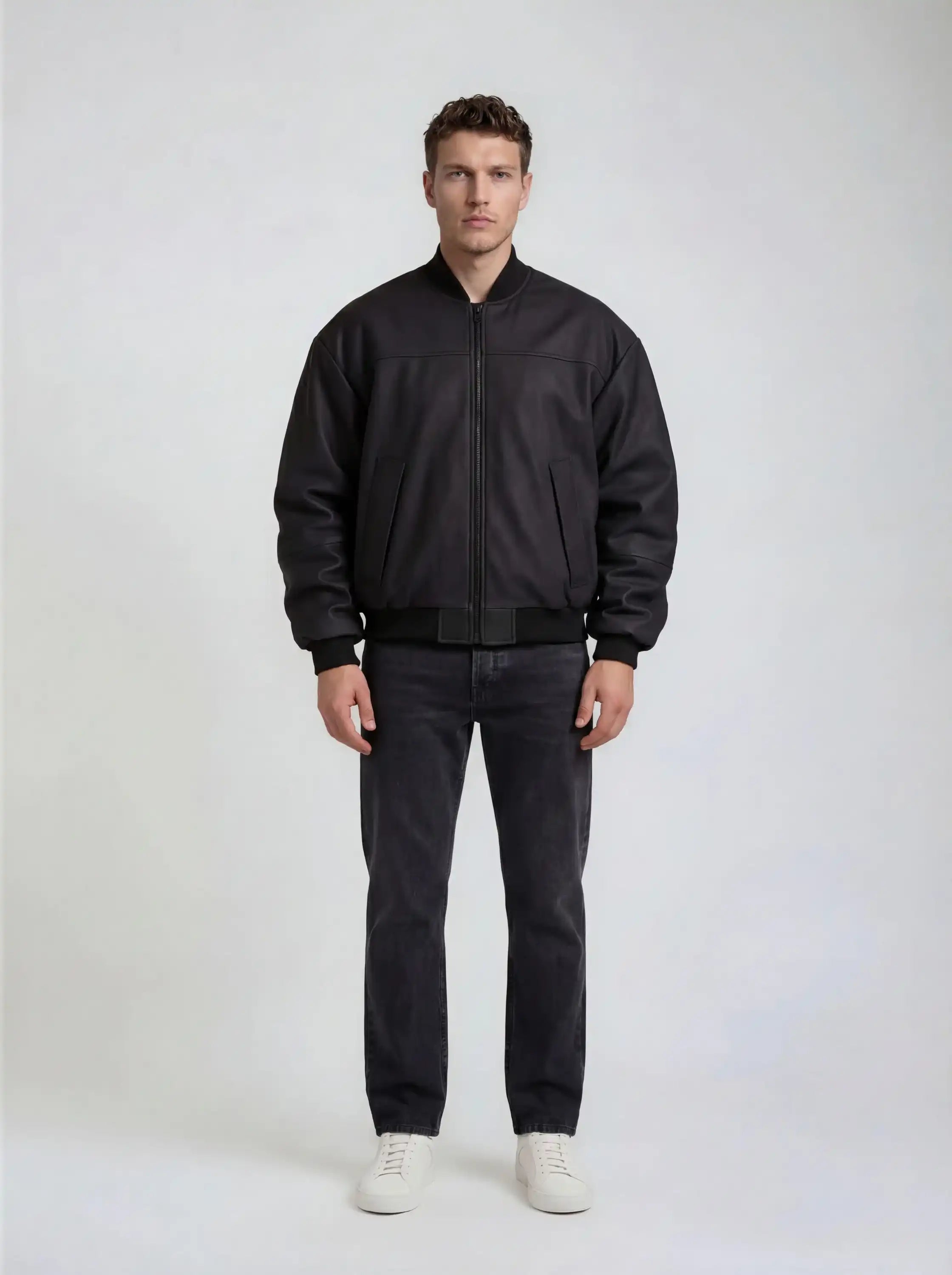 Ein Mann steht vor einem schlichten, hellen Hintergrund und trägt die trueprodigy Bomber Lederjacke Herren "Nazar" im Oversize-Schnitt, gepaart mit einer dunklen Jeans und weißen Sneakers. Seine Arme hängen entspannt an seinen Seiten und sein Gesichtsausdruck ist neutral.