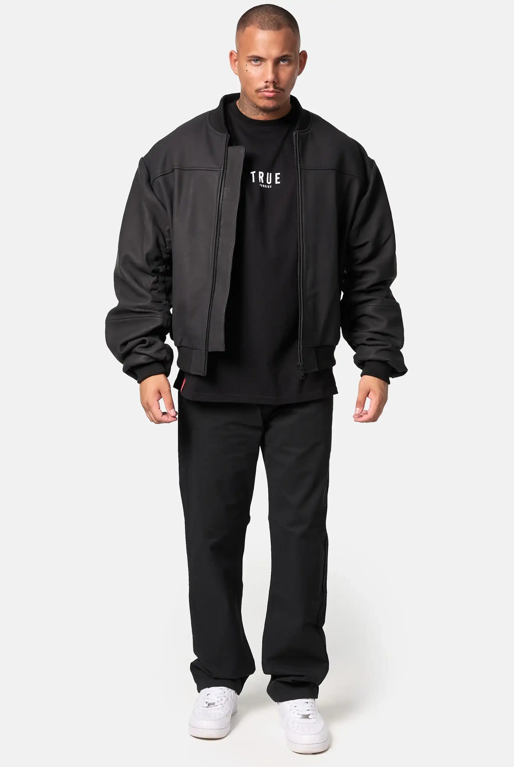 Ein Mann steht mit dem Gesicht nach vorne und trägt eine matte Trueprodigy Nazar Oversized Bomber-Lederjacke, ein schwarzes T-Shirt mit weißem Text, eine schwarze Hose und weiße Turnschuhe vor einem schlichten hellen Hintergrund.