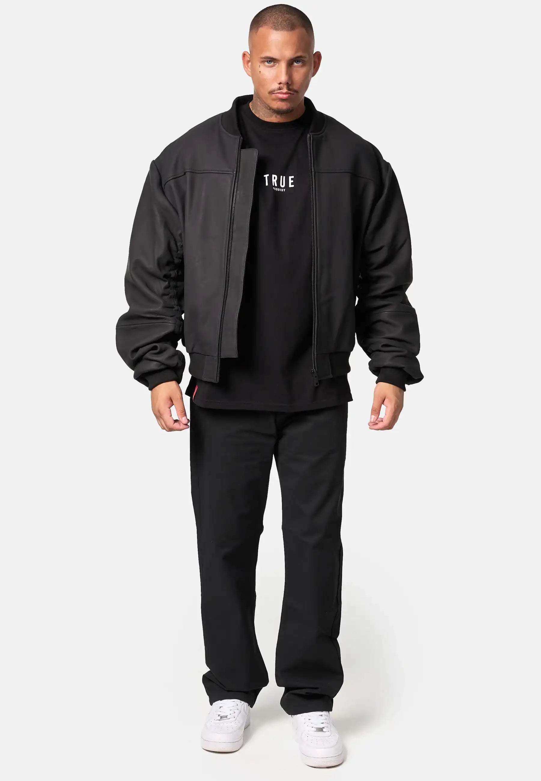 Ein Mann steht mit dem Gesicht nach vorne und trägt eine matte Trueprodigy Nazar Oversized Bomber-Lederjacke, ein schwarzes T-Shirt mit weißem Text, eine schwarze Hose und weiße Turnschuhe vor einem schlichten hellen Hintergrund.