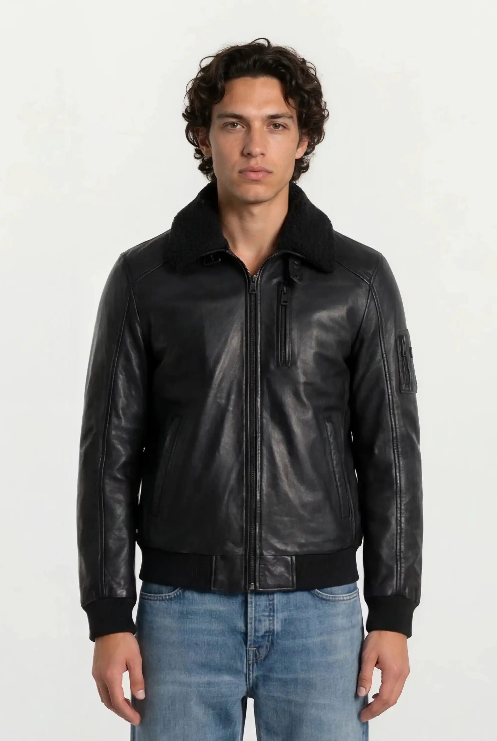 Eine Person mit mittellangem, lockigem Haar trägt eine trueprodigy Moderne Fliegerjacke aus echtem Leder mit abnehmbarem Teddy-Kragen und hellblaue Jeans vor einem schlichten hellen Hintergrund.