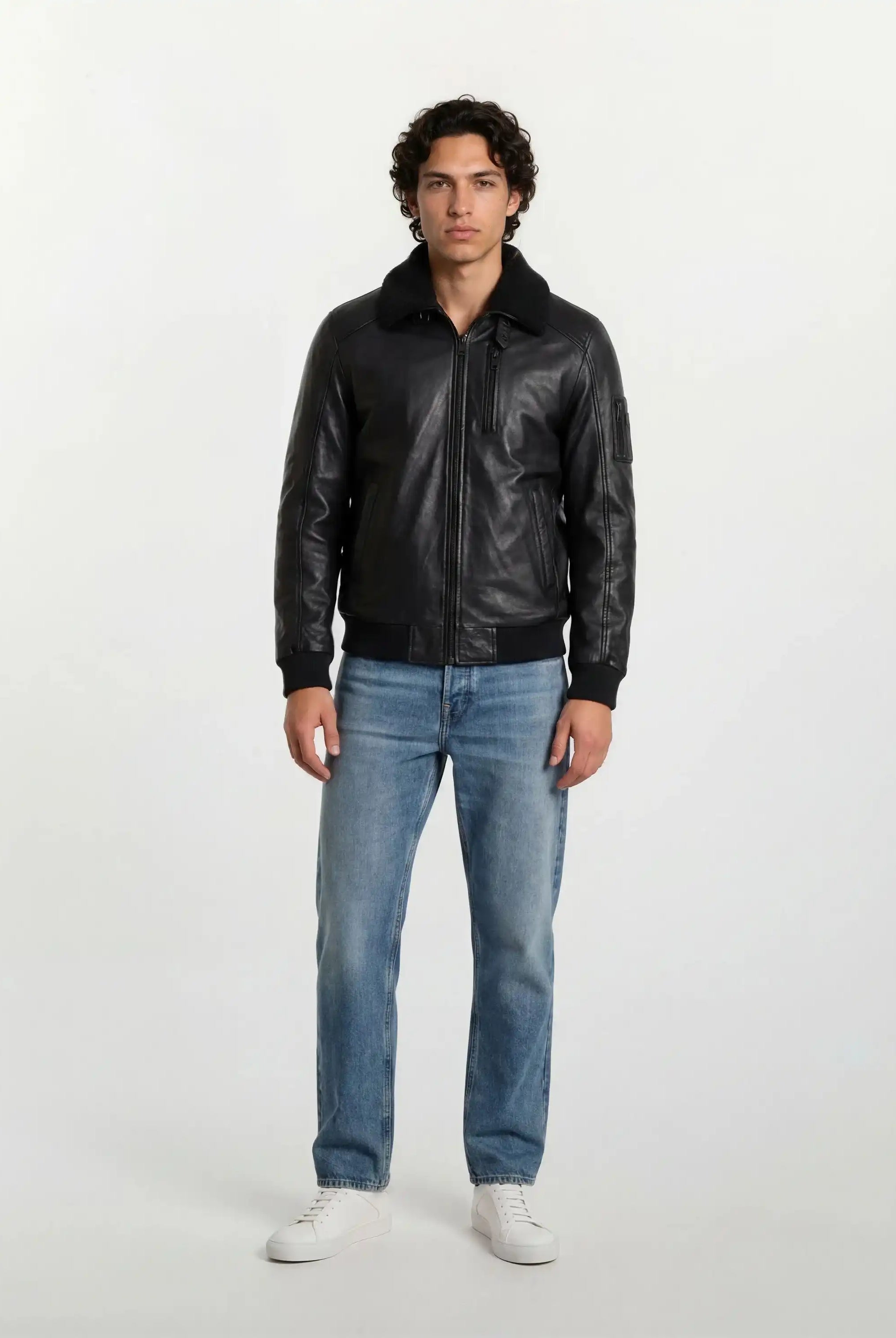 Ein Mann mit kurzen, lockigen dunklen Haaren steht mit dem Gesicht nach vorne vor einem weißen Hintergrund und trägt die trueprodigy Moderne Fliegerjacke aus echtem Leder mit abnehmbarem Teddy-Kragen, hellblaue Jeans und weiße Turnschuhe. Seine Arme ruhen an den Seiten.