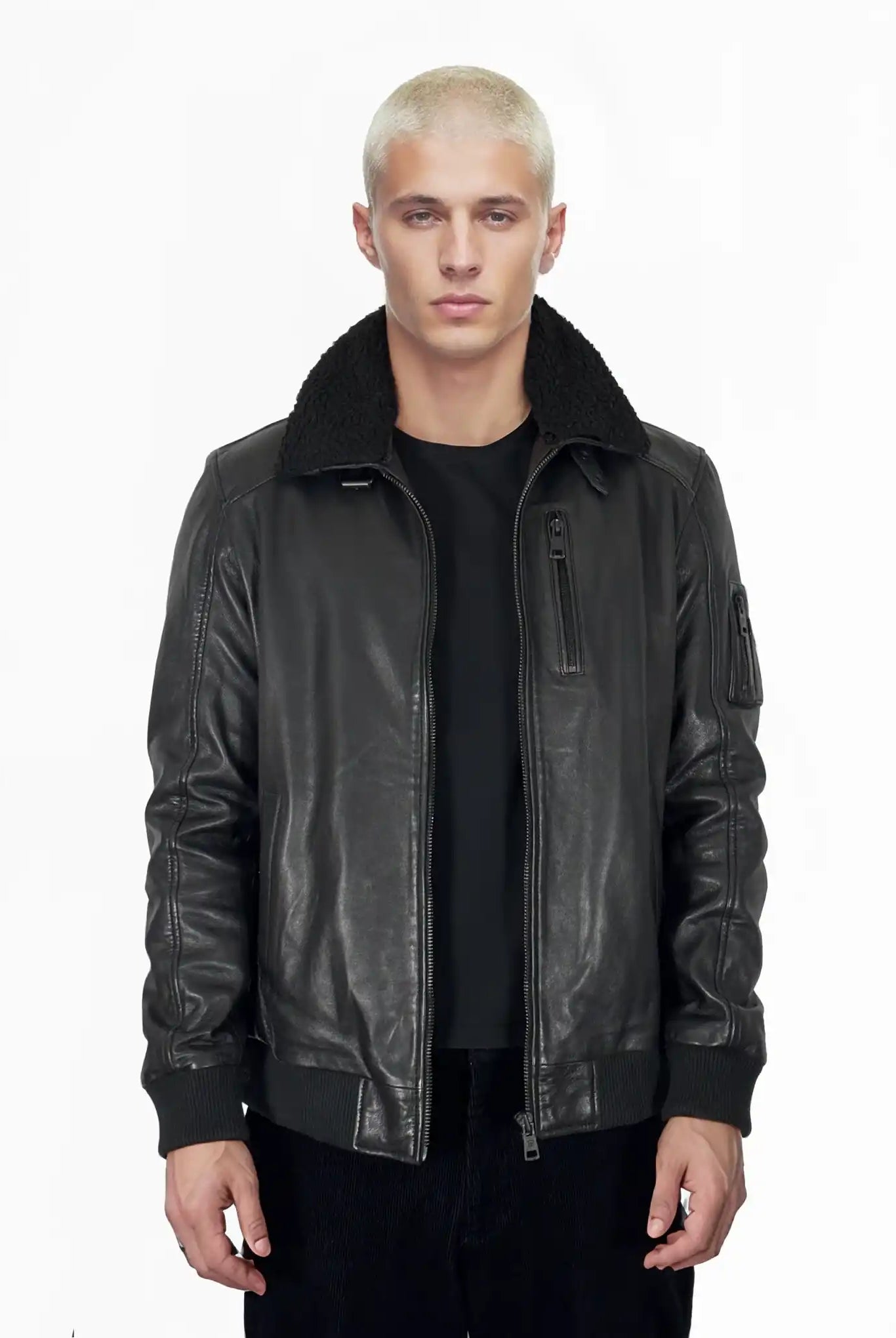 Eine Person mit kurzen, gebleichten blonden Haaren trägt die trueprodigy Blaze Moderne Fliegerjacke aus echtem Leder mit abnehmbarem Teddy-Kragen und ein schwarzes Hemd, stehend vor einem schlichten weißen Hintergrund.