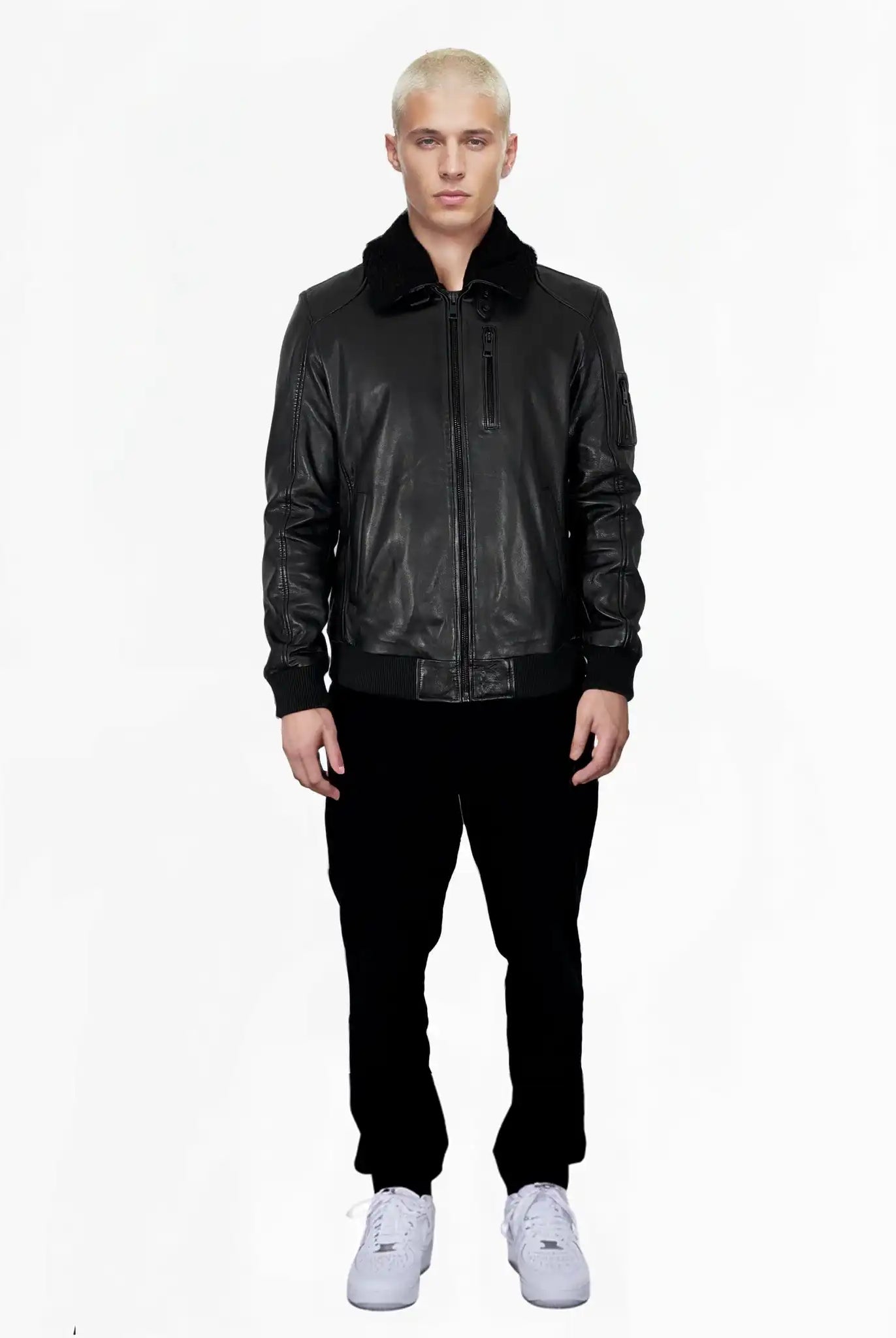Eine Person mit kurzen blonden Haaren steht vor der Kamera und trägt die Blaze Moderne Fliegerjacke aus echtem Leder mit abnehmbarem Teddy-Kragen von trueprodigy, schwarze Hosen und weiße Sneaker vor einem schlichten weißen Hintergrund.
